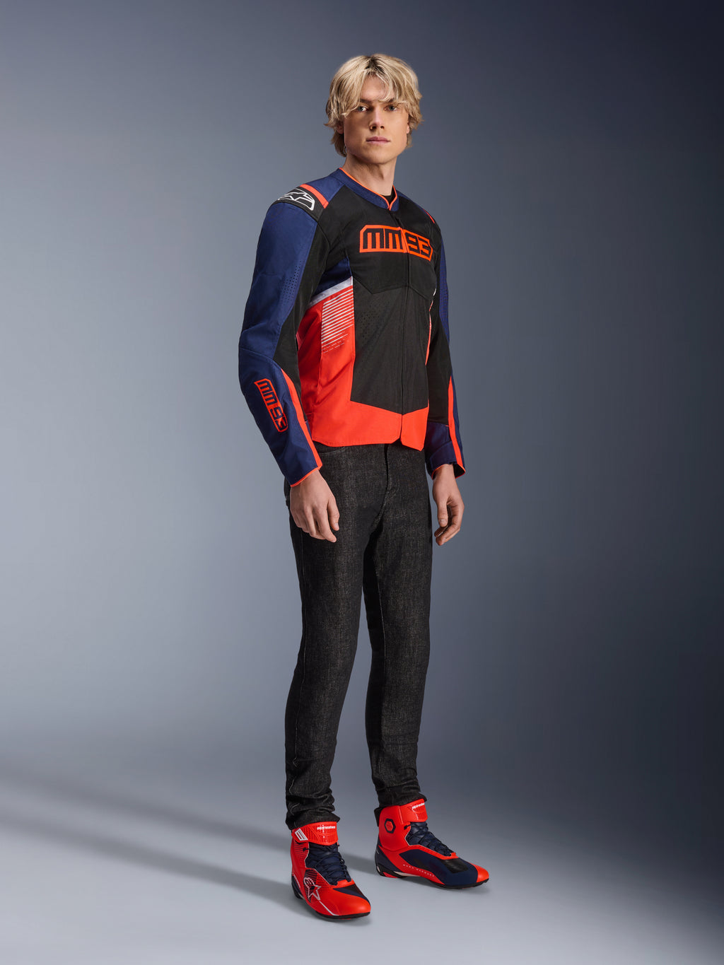 Modelo masculino vistiendo la chaqueta Alpinestars MM93 T-SPS Air V2 en azul marino, negro y rojo flúor, con el distintivo logotipo MM93 en el pecho y las mangas, combinada con vaqueros oscuros y botas de moto rojas y azules a juego, equipamiento deportivo de conducción urbana