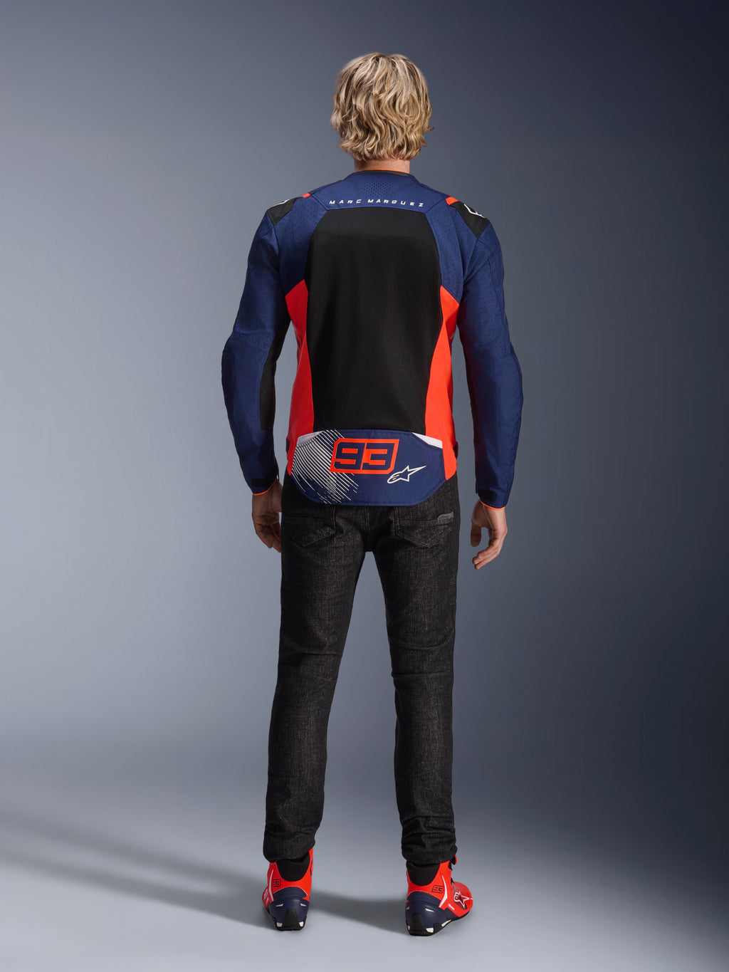 Vista posterior de un modelo con la Chaqueta MM93 T-SPS Air V2 en azul marino con detalles en rojo flúor y negro, que cuenta con un gran panel de malla en la espalda y la marca '93' de Marc Márquez, combinada con pantalones denim oscuros y botas de motociclista a juego