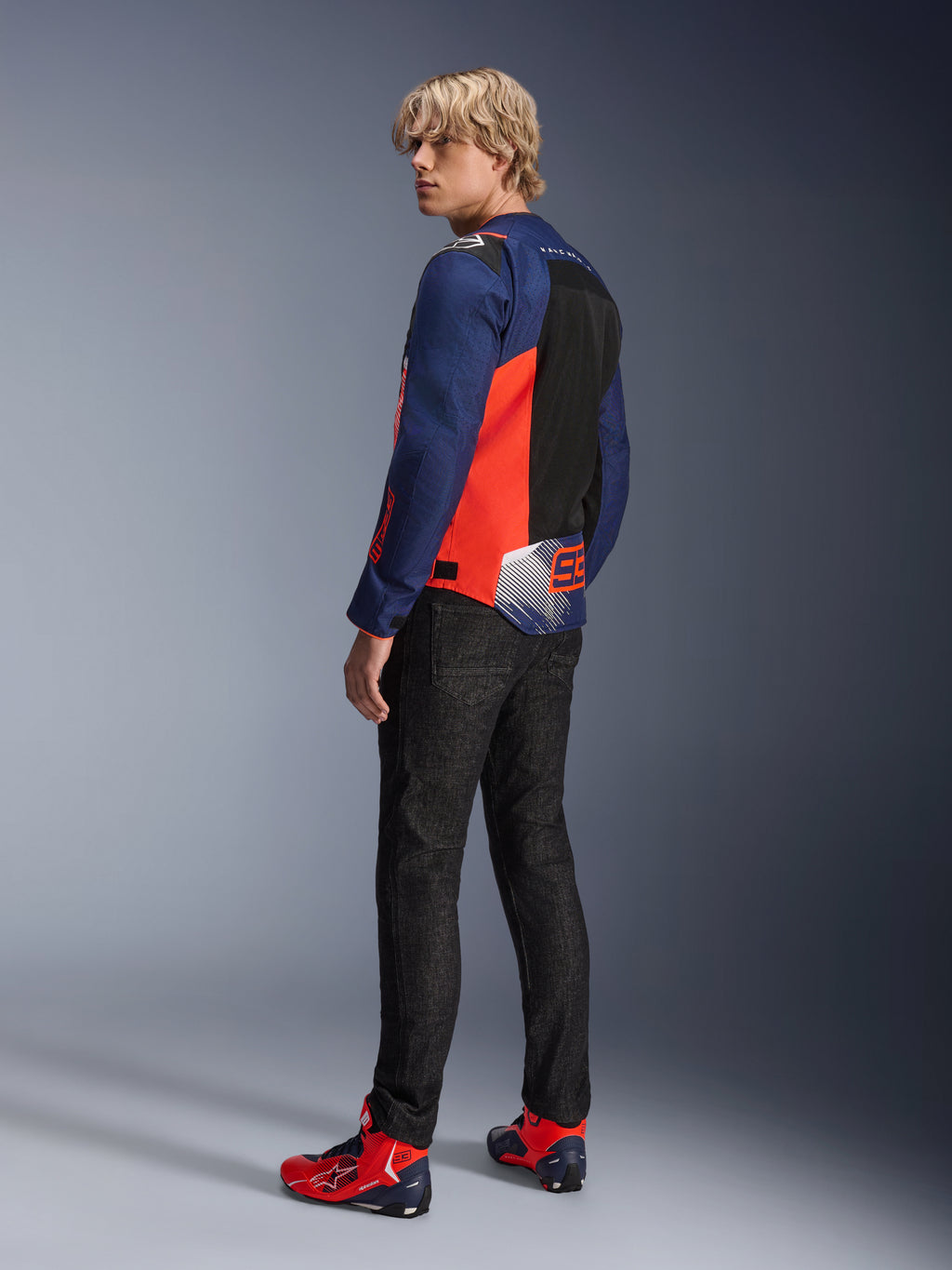 Alpinestars MM93 T-SPS Air V2 Jacket, chaqueta de moto en azul marino y rojo flúor, vista trasera de un modelo con vaqueros negros y calzado rojo y azul a juego, con paneles de malla y logotipos de Marc Márquez 93