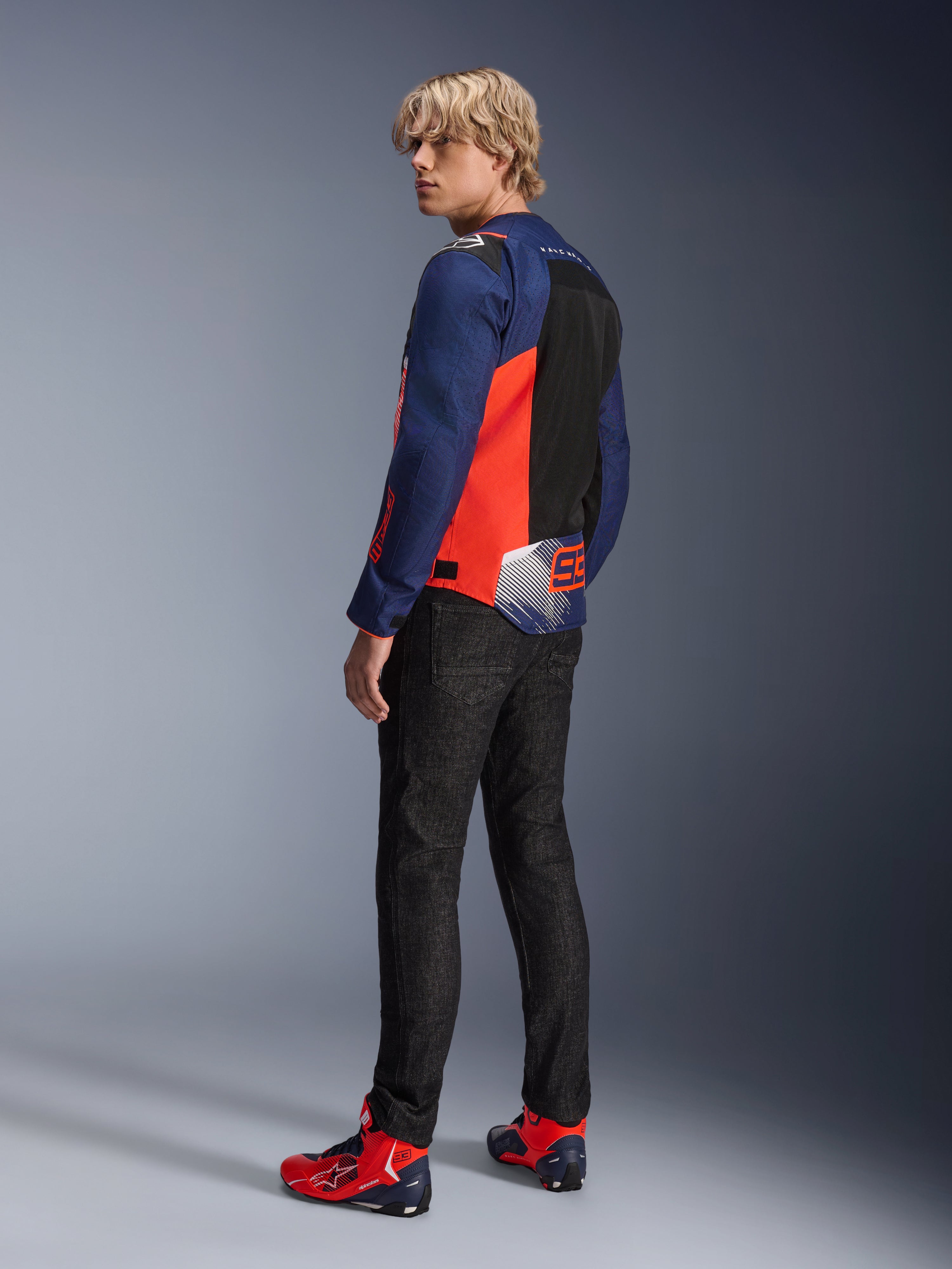 Alpinestars MM93 T-SPS Air V2 Jacket, chaqueta de moto en azul marino y rojo flúor, vista trasera de un modelo con vaqueros negros y calzado rojo y azul a juego, con paneles de malla y logotipos de Marc Márquez 93