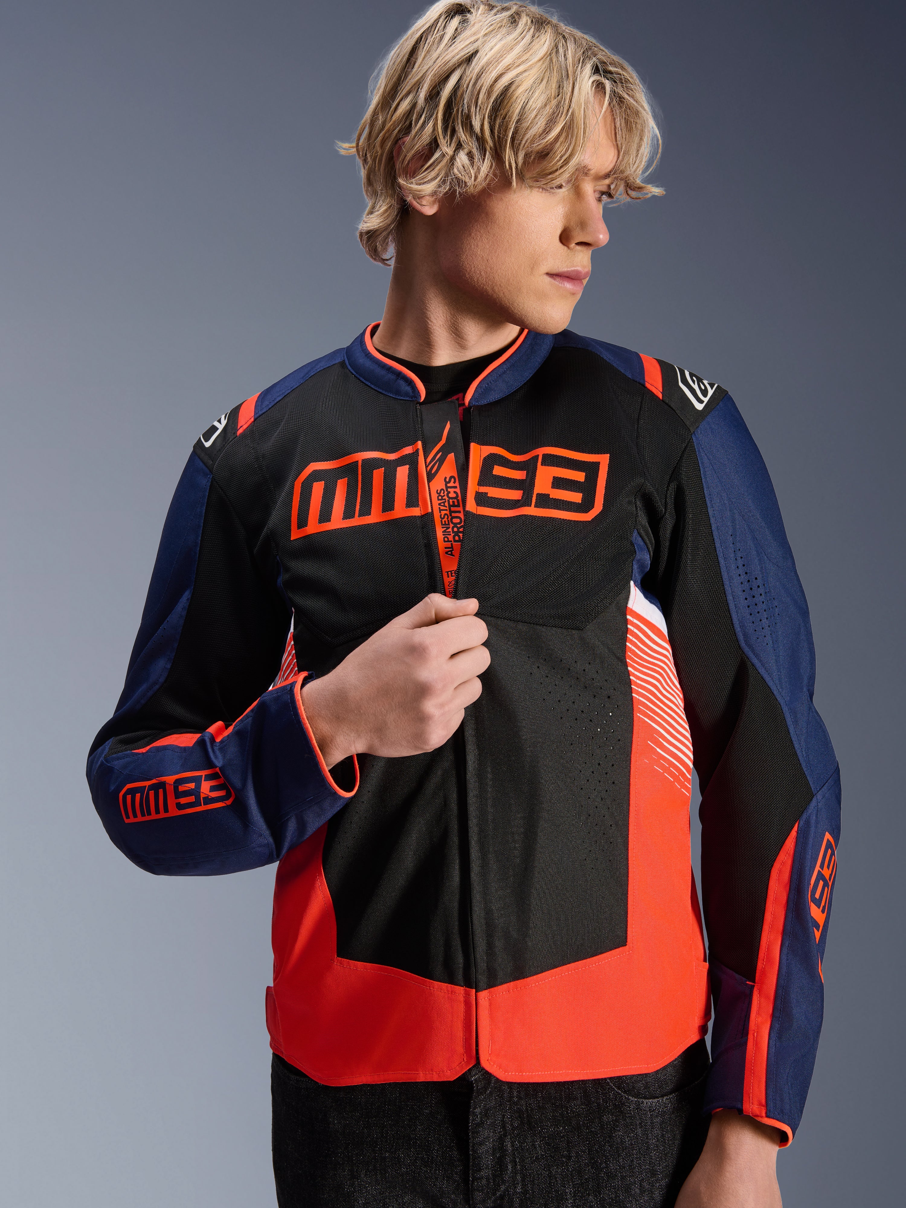 Chaqueta Alpinestars MM93 T-SPS Air V2, chaqueta de moto textil en azul marino, negro y rojo fluorescente, usada por un modelo masculino que ajusta la cremallera, con llamativos logotipos de Marc Márquez MM93 en el pecho y las mangas, y paneles de malla ventilada para conducción en carretera durante el verano.