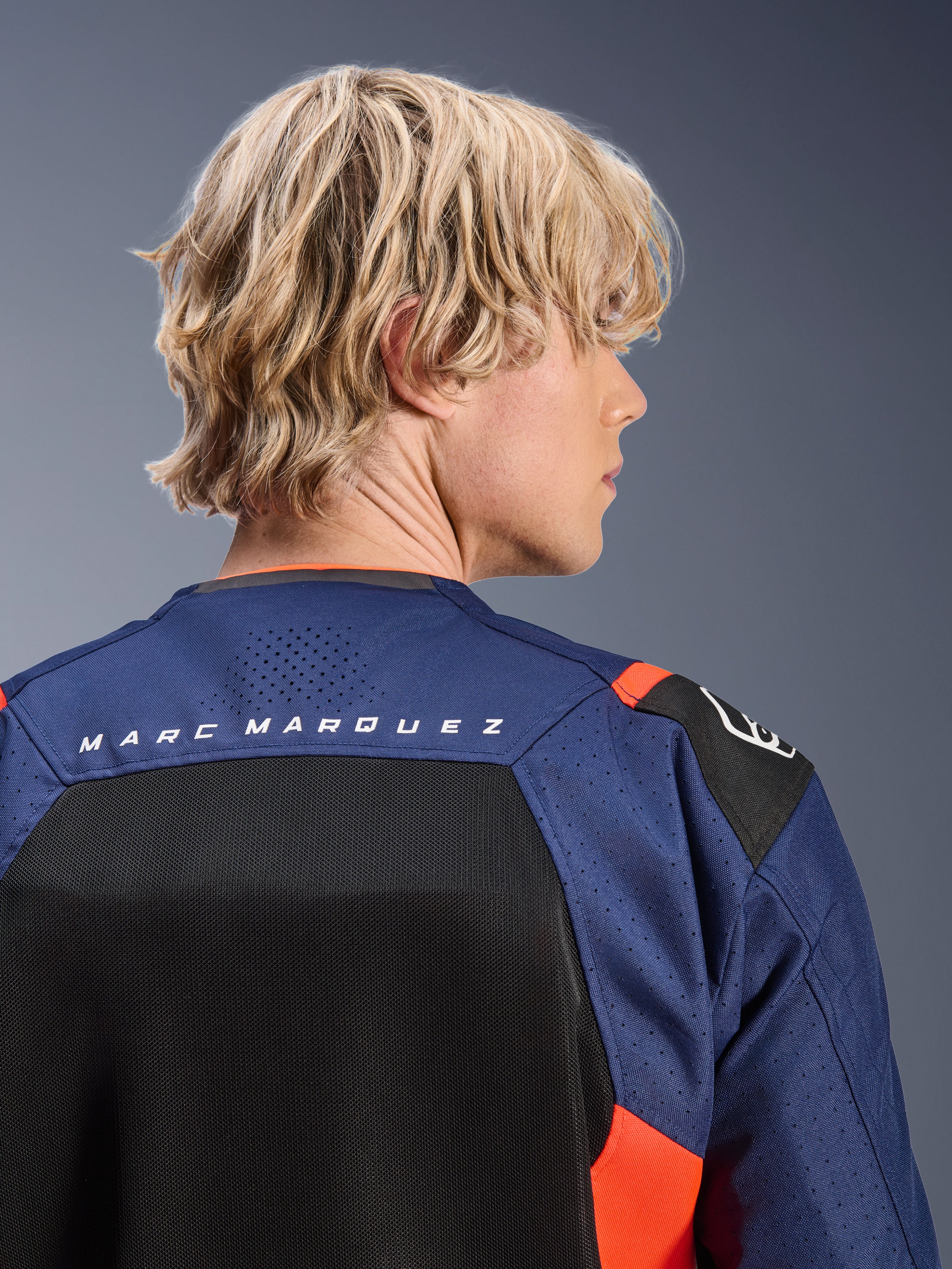 Chaqueta Alpinestars MM93 T-SPS Air V2, azul marino y negro con detalles en rojo flúor, primer plano de la parte superior de la espalda con la marca 'MARC MARQUEZ', paneles de malla transpirable y construcción textil perforada, usada por un modelo rubio.