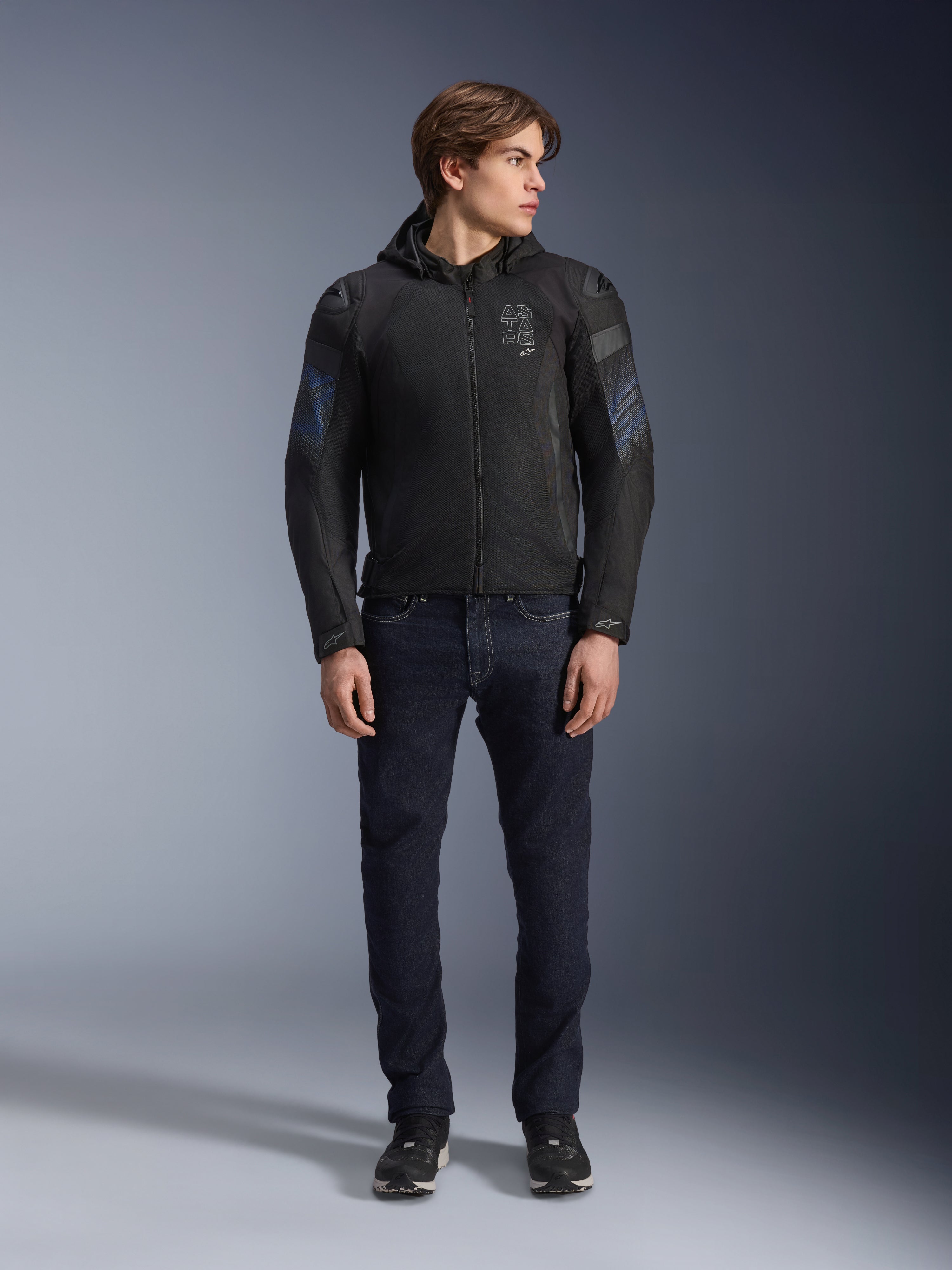 Modelo masculino con la chaqueta Alpinestars Zaca Air Venom WP en negro y azul eléctrico, chaqueta de moto textil con paneles de malla 3D y capucha desmontable, combinada con vaqueros de denim oscuro, equipamiento de conducción moderno de inspiración deportiva.