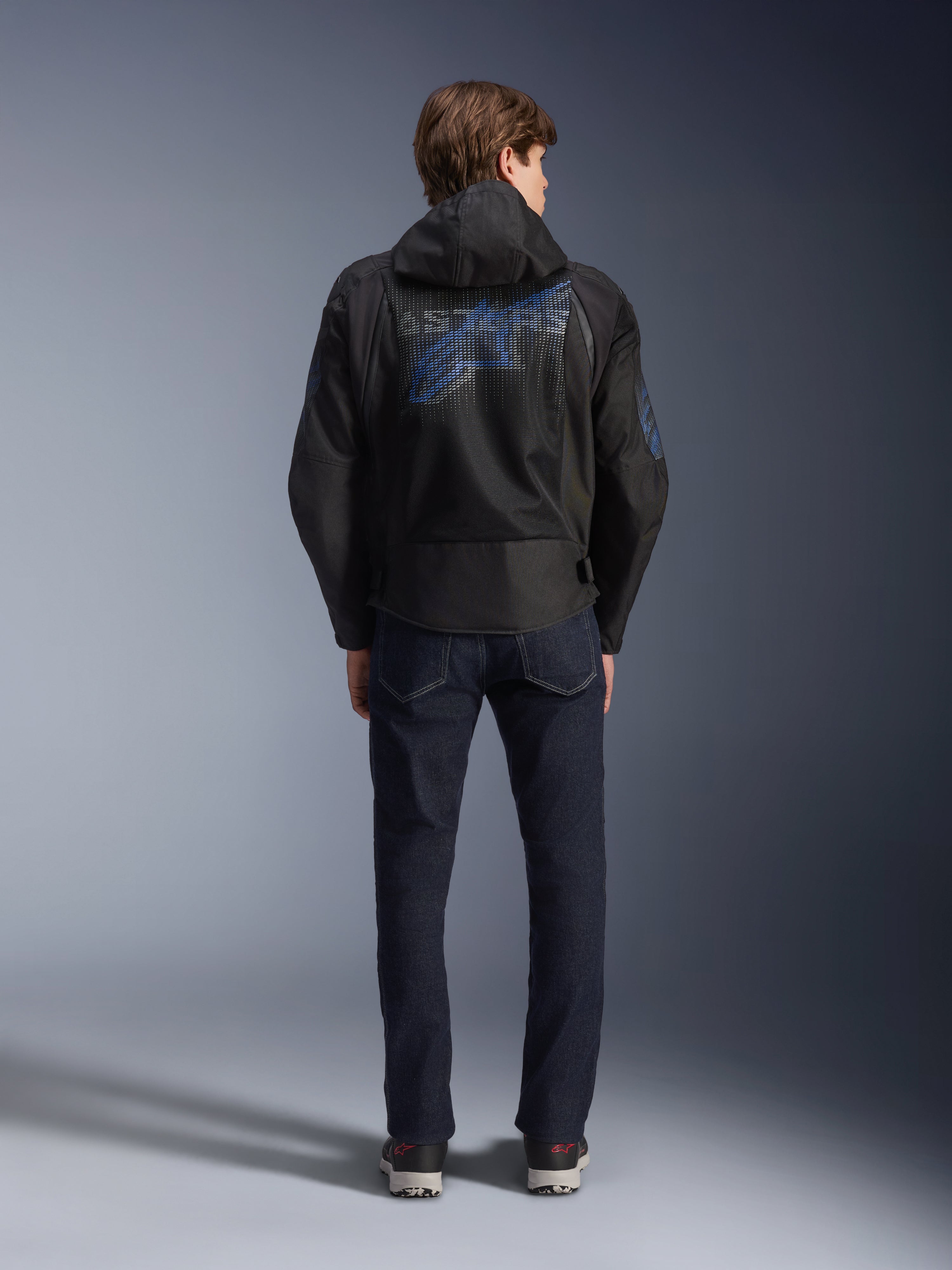 Chaqueta Alpinestars Zaca Air Venom WP, negro y azul eléctrico, vista posterior con un gran logotipo azul de Alpinestars en el panel de malla trasero, capucha desmontable, combinada con vaqueros de denim oscuro y zapatillas, equipamiento deportivo de moto para todo tipo de clima.