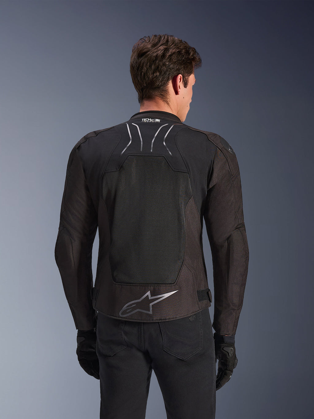 Chaqueta Alpinestars SMX Air en color Black Anthracite, vista posterior en un modelo, con un gran panel trasero de malla técnica para ventilación, logotipo Tech-Air Ready en el cuello y un logotipo Astar plateado en la parte inferior de la espalda, combinada con guantes negros y pantalones de montar.