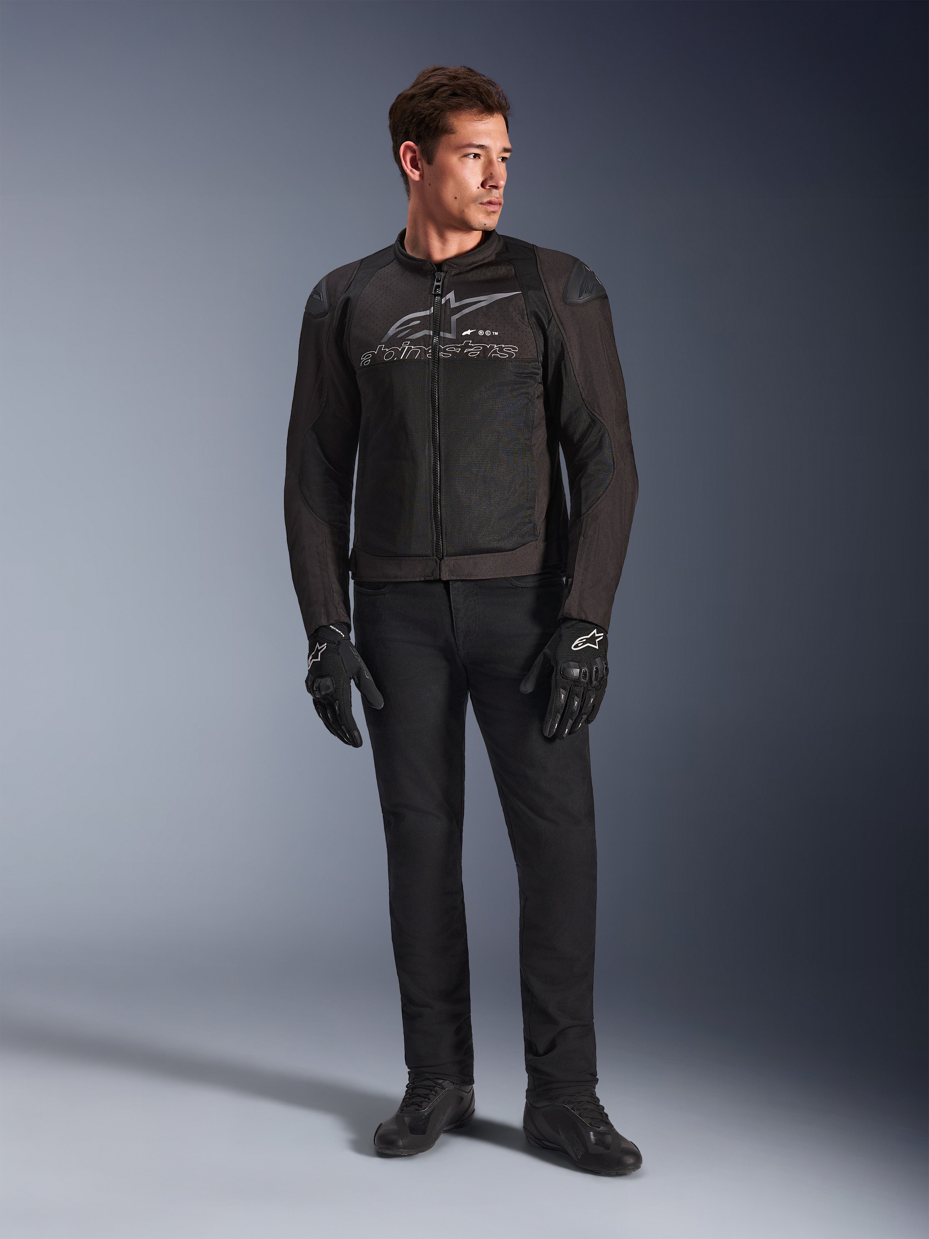 Modelo masculino con la chaqueta de moto Alpinestars SMX Air en color Black Anthracite, con paneles de malla ventilada y logo gris en el pecho, combinada con pantalones negros y guantes protectores, en un entorno de estudio con fondo degradado