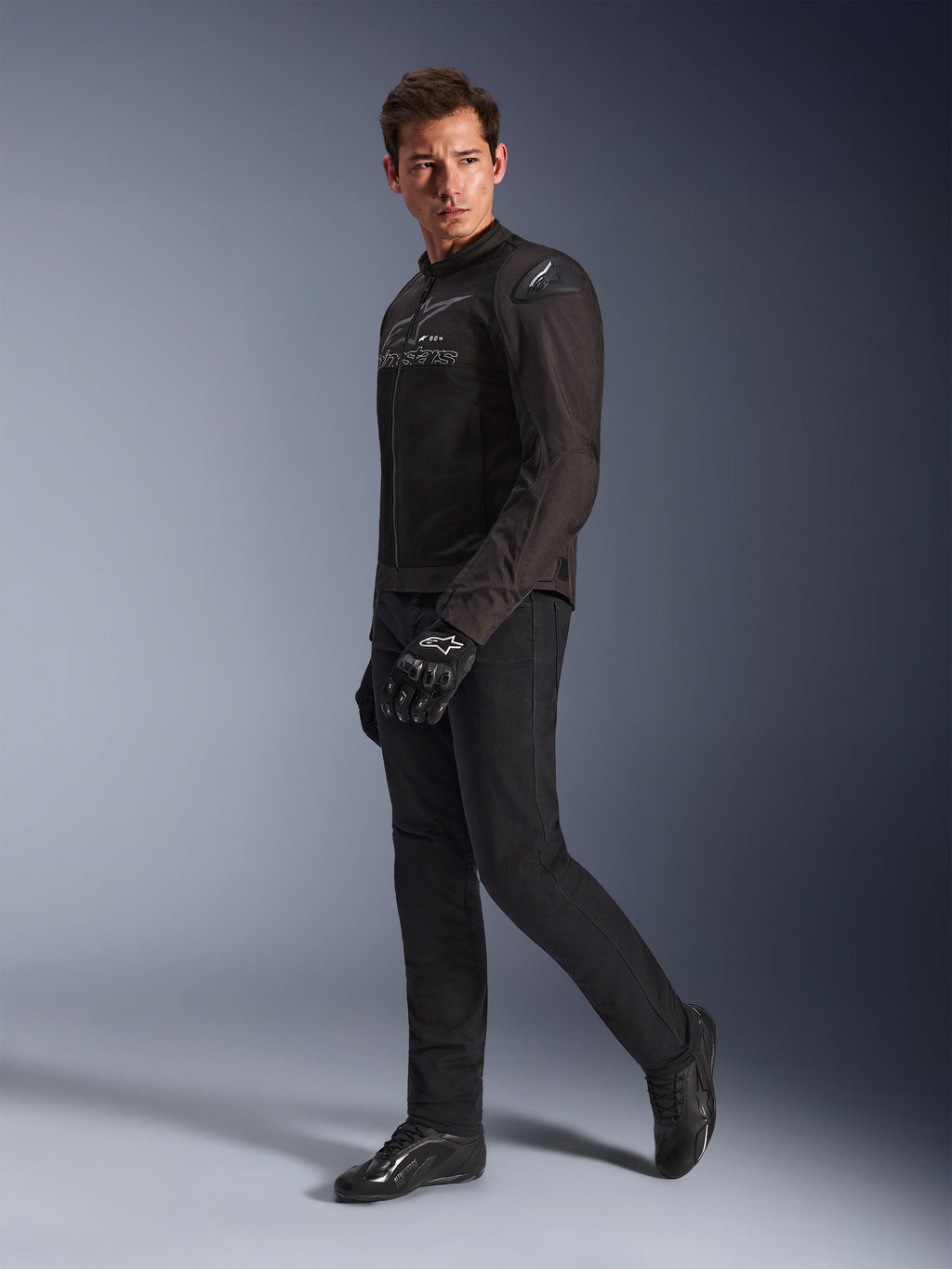 Chaqueta Alpinestars SMX Air en Negro Antracita, usada por un modelo con pantalones de motociclista, guantes y calzado negros a juego, toma de estudio de cuerpo completo, equipamiento deportivo de alto rendimiento para motociclismo con malla técnica y deslizadores de hombro