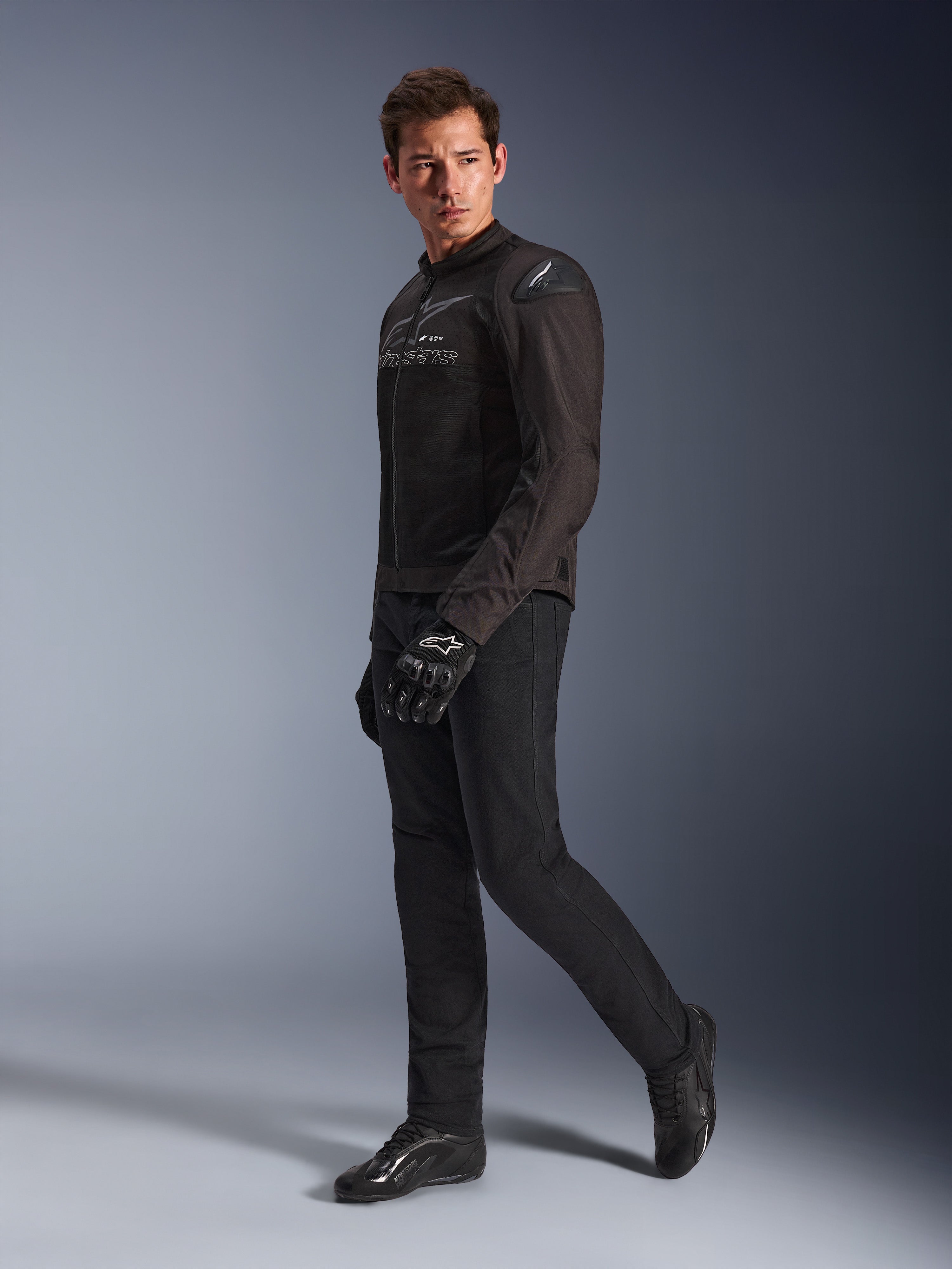 Chaqueta Alpinestars SMX Air en Negro Antracita, usada por un modelo con pantalones de motociclista, guantes y calzado negros a juego, toma de estudio de cuerpo completo, equipamiento deportivo de alto rendimiento para motociclismo con malla técnica y deslizadores de hombro