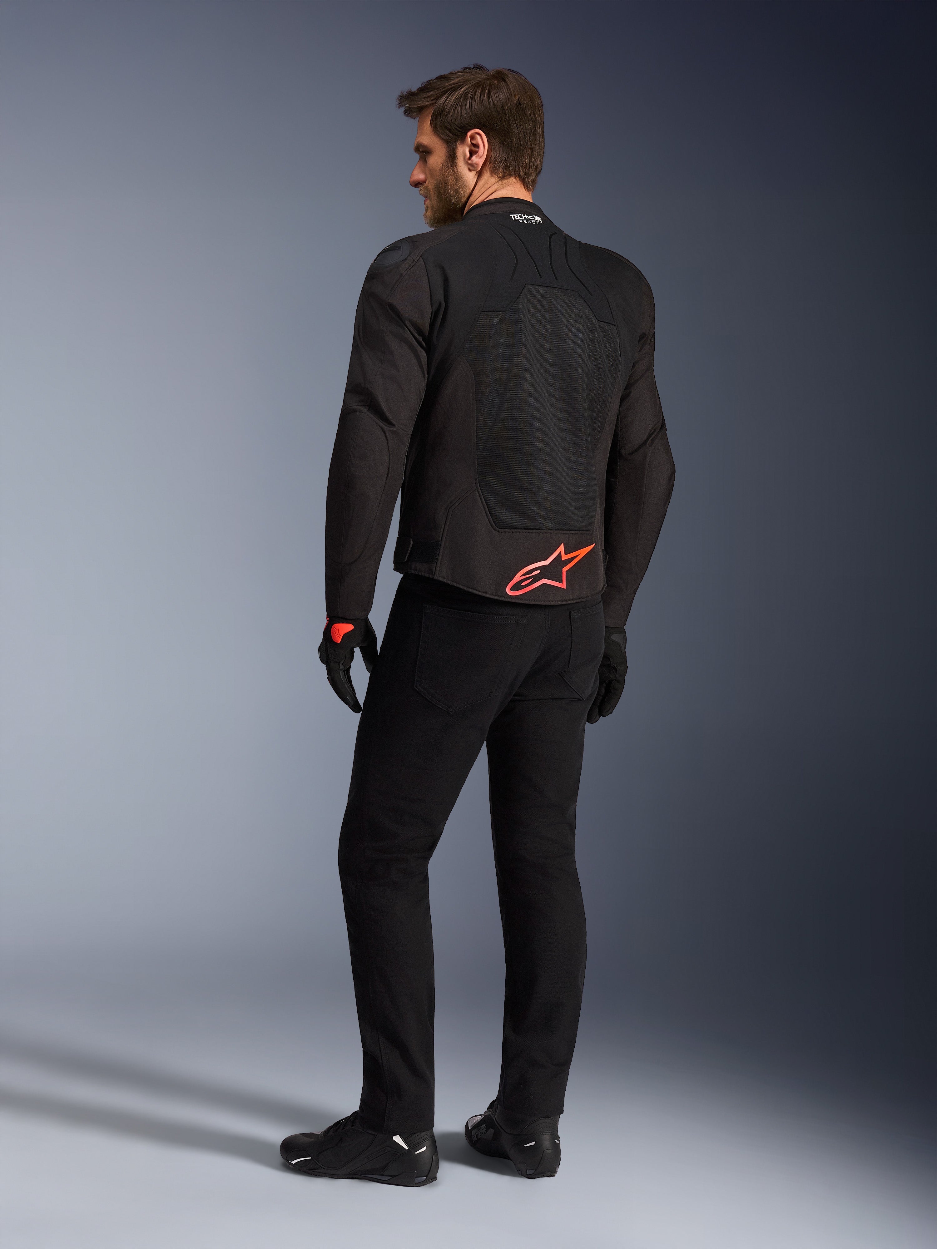 Vista trasera de un modelo luciendo la chaqueta Alpinestars SMX Air en negro, con un logotipo de Alpinestars en rojo brillante en la parte inferior de la espalda, la marca 'Tech-Air Ready' en el cuello y paneles de malla técnica para ventilación, combinada con pantalones y guantes de conducción negros.