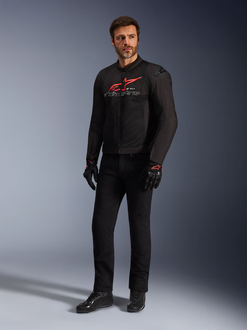 Modelo masculino vistiendo la chaqueta Alpinestars SMX Air en negro, rojo brillante y blanco, combinada con pantalones técnicos negros, guantes negros y rojos y calzado de conducción negro, con un logotipo rojo destacado en el pecho y detalles de marca en blanco, toma de estudio de cuerpo completo para equipamiento de motociclismo deportivo