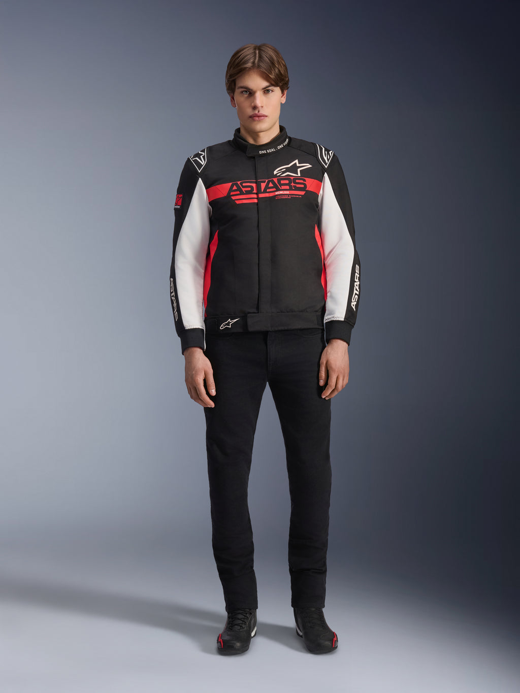 Male model wearing the Alpinestars Monza Sport Jacket in Black, Bright Red, and White, featuring a large red 'ASTARS' logo across the chest and white panels on the sleeves, paired with black pants and black riding boots.