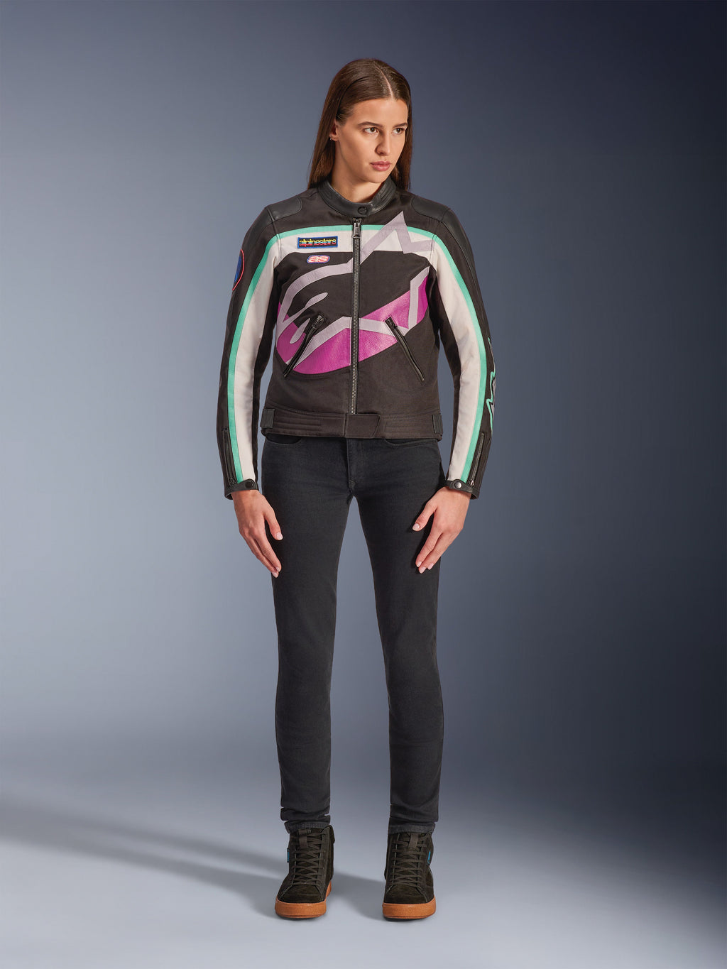 Woman wearing Alpinestars Idles Women’s Jacket, Black Off-White and Purple, vintage racing-inspired design with large wrap-around logo and teal accents, styled with black jeans and riding sneakers, urban motorcycle apparel