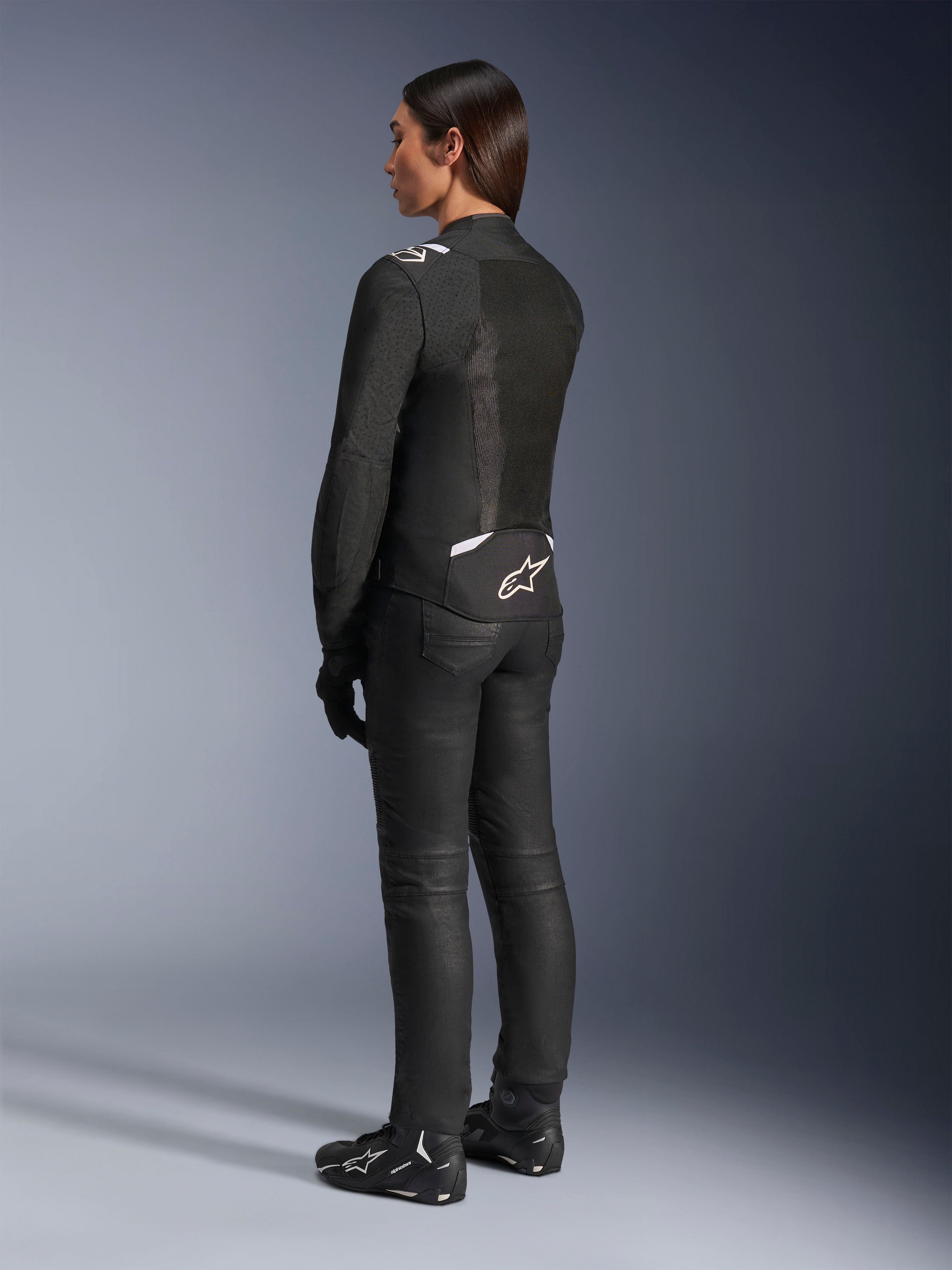 Chaqueta Alpinestars Stella T-Sps Air V2, negra y blanca, vista trasera que muestra paneles de malla y el logotipo en la parte inferior de la espalda, combinada con pantalones de conducción negros y calzado para moto, equipamiento textil deportivo de carretera para mujer