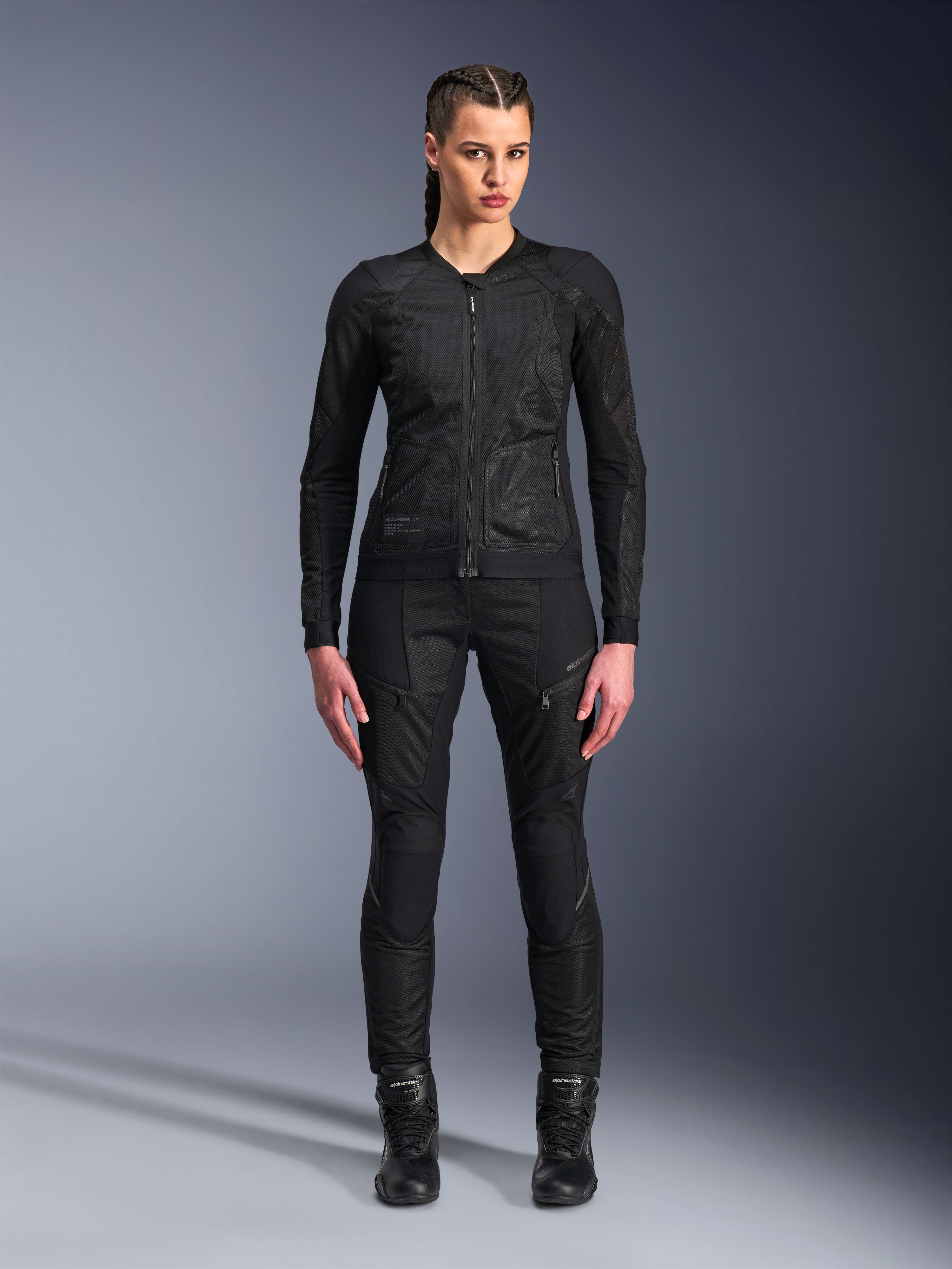 Chaqueta Alpinestars Stella Troop-Air, chaqueta de moto para mujer, negra, construcción de malla técnica 2D con diseño minimalista, usada con pantalones de conducción y botas negras, estilo de motociclismo urbano contemporáneo.