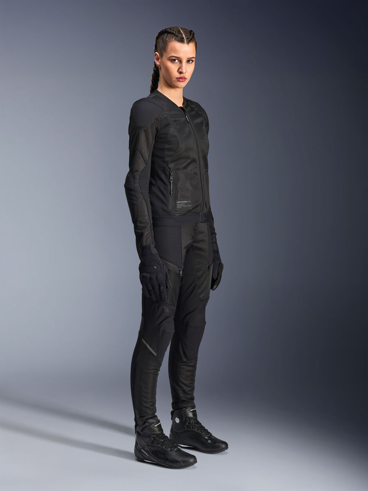 Mujer piloto vistiendo la chaqueta Alpinestars Stella Troop-Air en color negro, con construcción técnica de malla 2D, combinada con pantalones, guantes y botas de moto negros a juego, diseñada para conducción urbana