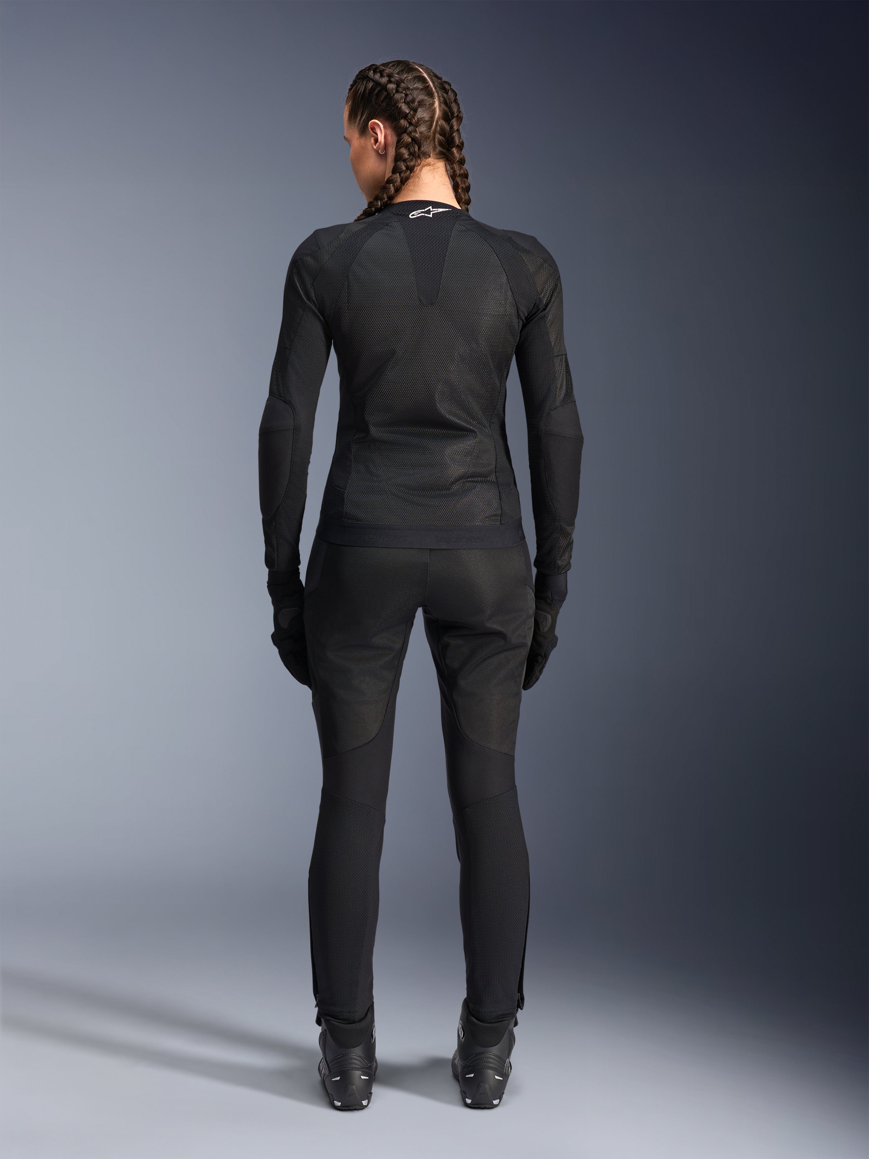 Chaqueta Alpinestars Stella Troop-Air, chaqueta de moto para mujer, negra, vista trasera de una piloto con chaqueta de malla técnica, combinada con pantalones de montar, guantes y botas negros a juego, diseño transpirable de ajuste ceñido con un logotipo blanco de Alpinestars en el cuello