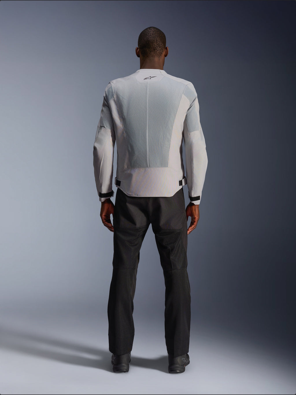 Chaqueta Alpinestars C-1 Air en color plata, vista desde atrás en un modelo masculino, chaqueta de moto de tejido técnico con amplios paneles de ventilación de malla técnica, pequeño logotipo negro en el cuello y ajustadores de cintura, combinada con pantalones de conducción negros para un estilo urbano contemporáneo.