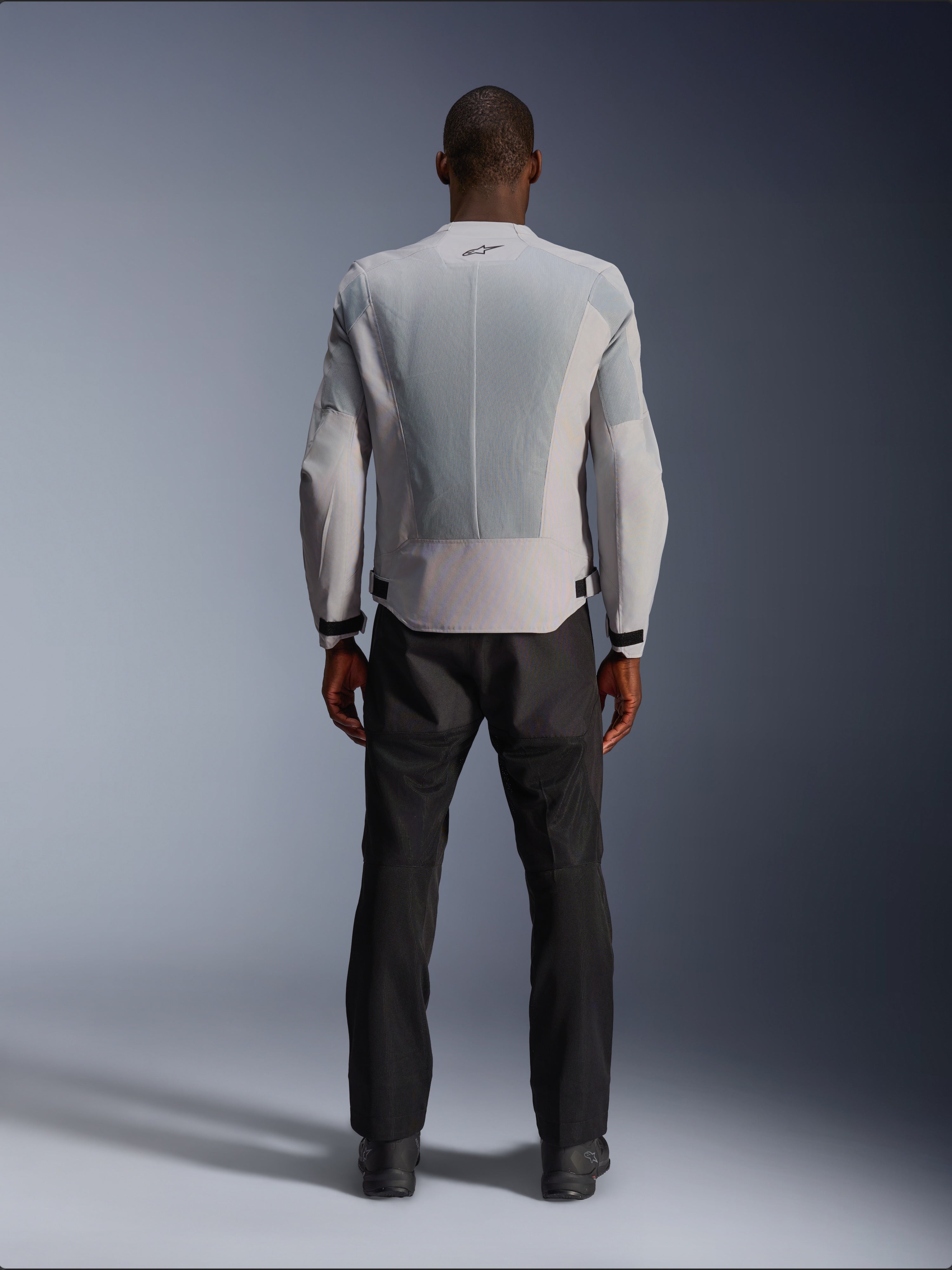 Chaqueta Alpinestars C-1 Air en color plata, vista desde atrás en un modelo masculino, chaqueta de moto de tejido técnico con amplios paneles de ventilación de malla técnica, pequeño logotipo negro en el cuello y ajustadores de cintura, combinada con pantalones de conducción negros para un estilo urbano contemporáneo.
