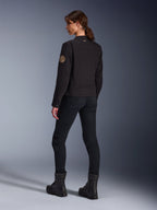 Modelo luciendo la chaqueta Stella Grace de Alpinestars en negro, vista posterior que destaca el parche espiral vintage Oscar en el brazo y el pequeño logo de estrella en el cuello, combinada con pantalones de moto negros y botas negras para conducción urbana