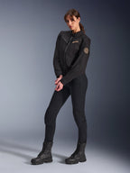 Modelo femenina con la chaqueta Stella Grace de Alpinestars para mujer, chaqueta urbana de moto negra, combinada con pantalones y botas de montar negros, diseño inspirado en la ropa de trabajo con logotipo bordado en bronce en el pecho y parche circular en espiral en la manga
