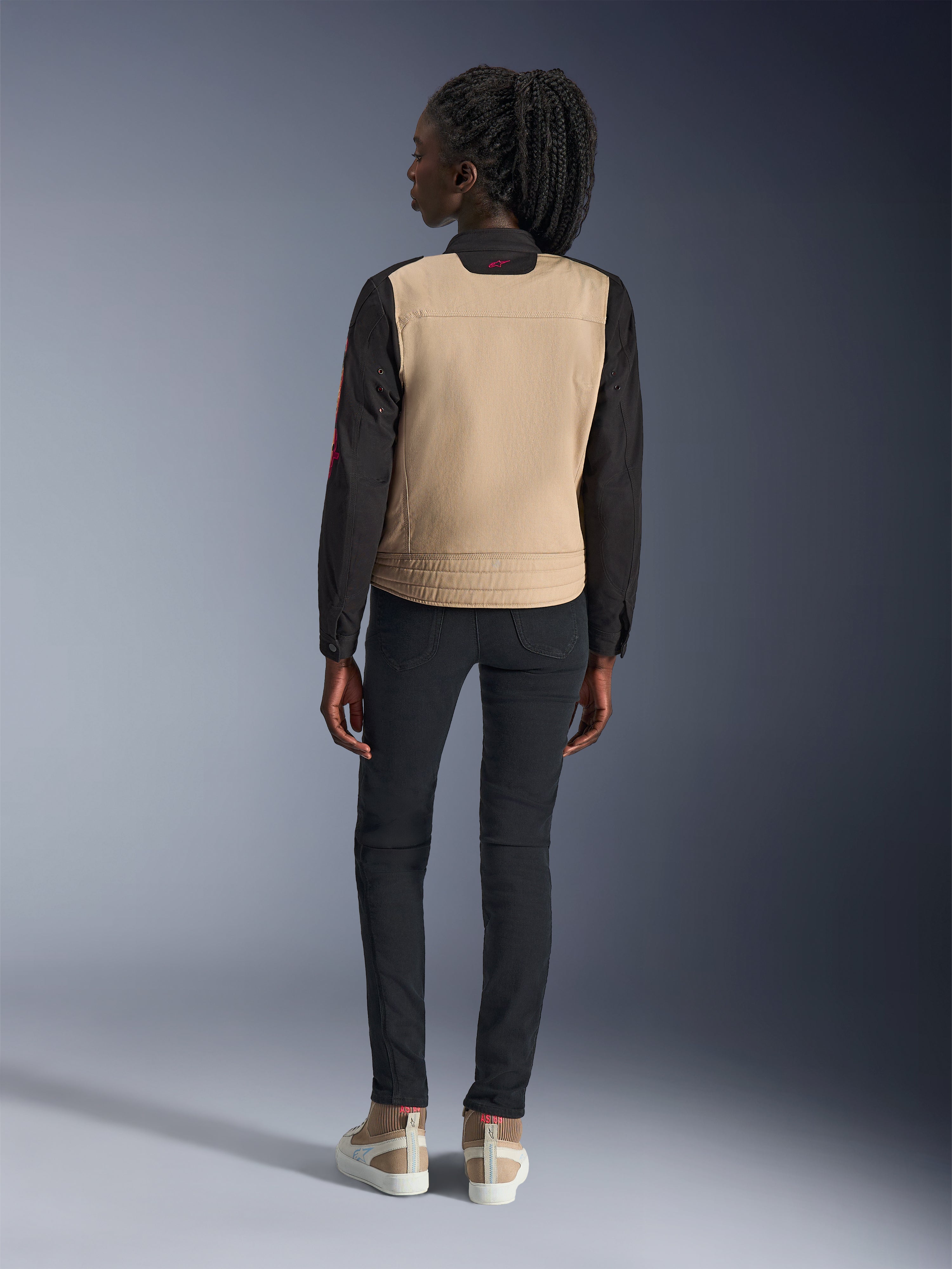 Mujer con chaqueta Alpinestars Stella Grace, color Aluminum Black con torso canela y mangas negras, vista trasera, combinada con pantalones ajustados negros y zapatillas casuales, equipamiento urbano de moto para mujer