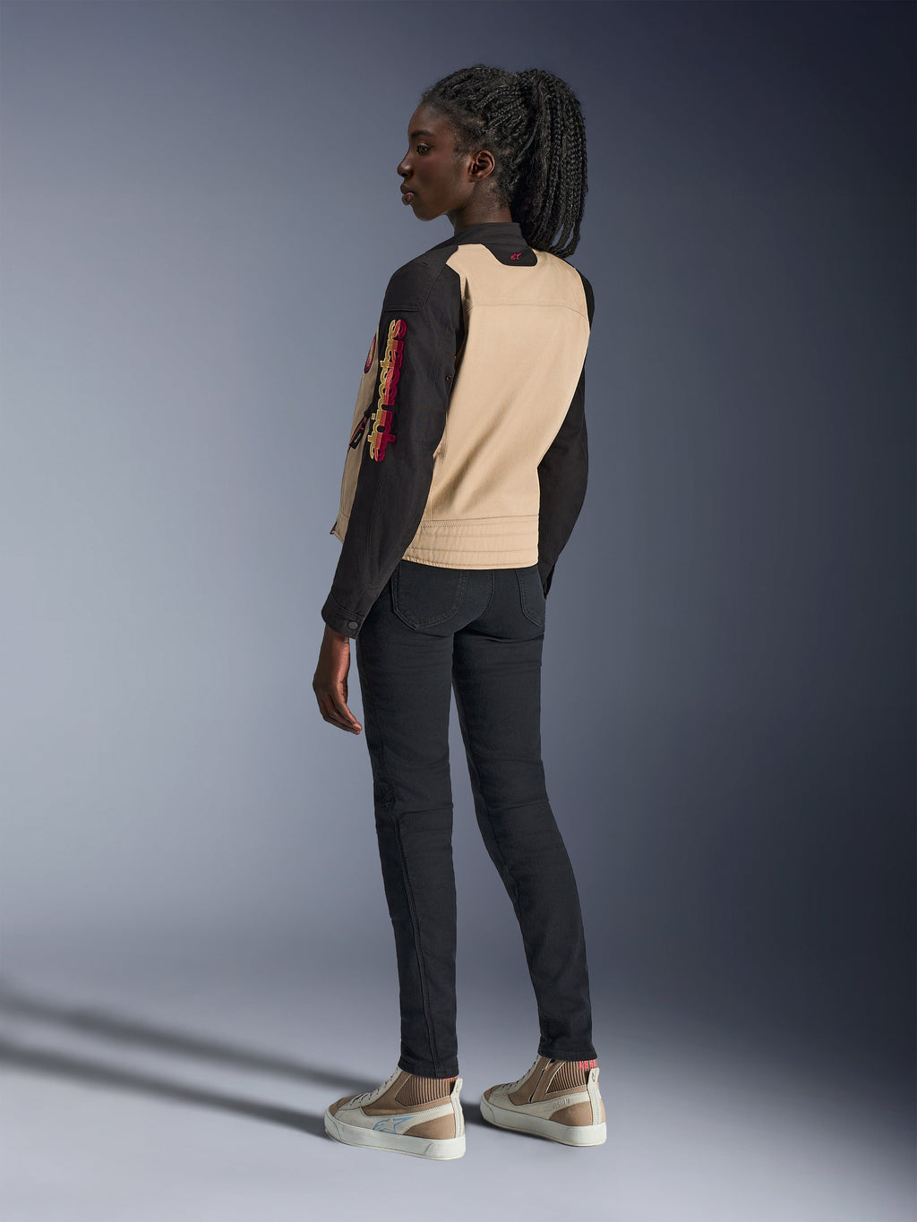 Chaqueta Alpinestars Stella Grace para mujer, chaqueta de moto urbana, negro aluminio con panel trasero canela y mangas negras, vista lateral trasera en una modelo, con logotipos coloridos bordados en la manga y construcción de hombros plisada