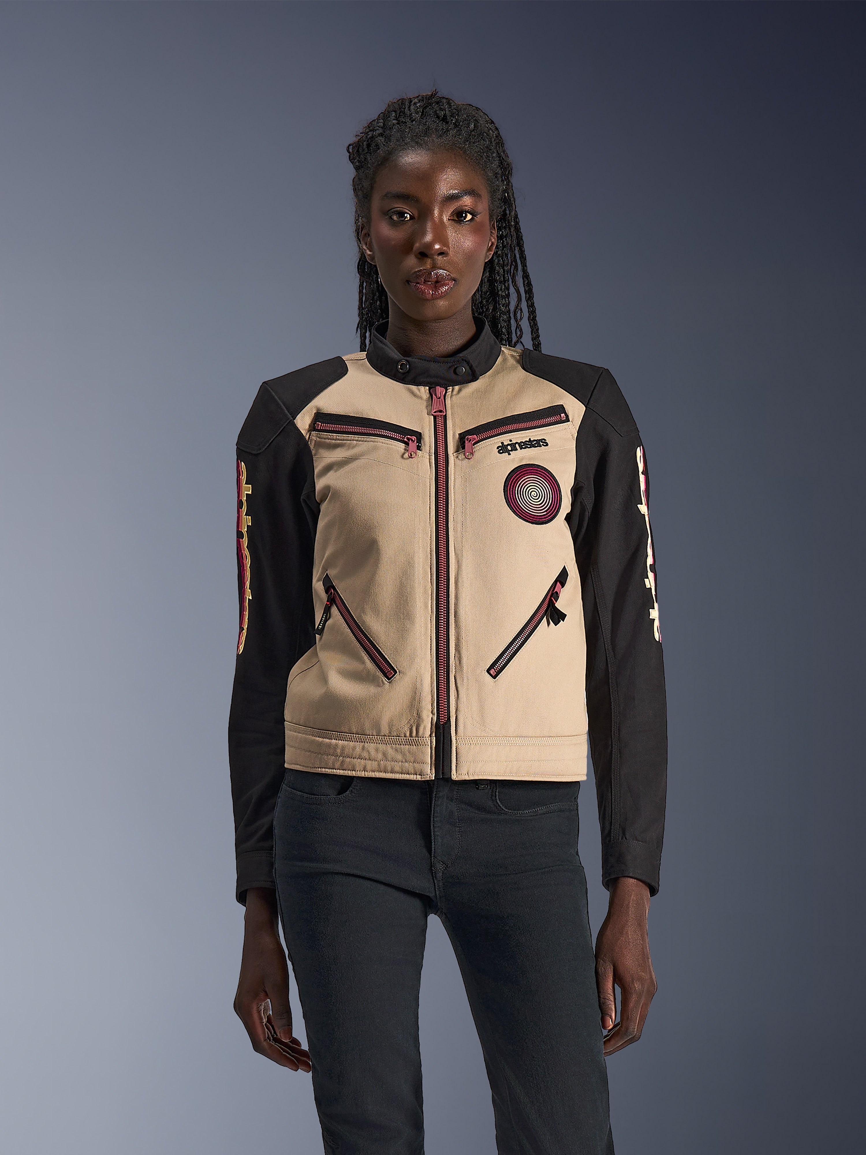 Alpinestars Stella Grace Jacket, chaqueta de moto para mujer, aluminio y negro, con un diseño inspirado en la ropa de trabajo, detalles de cremalleras en rojo rosado, un parche de espiral en el pecho y mangas largas negras con gráficos amarillos y rojos, usada por una modelo.
