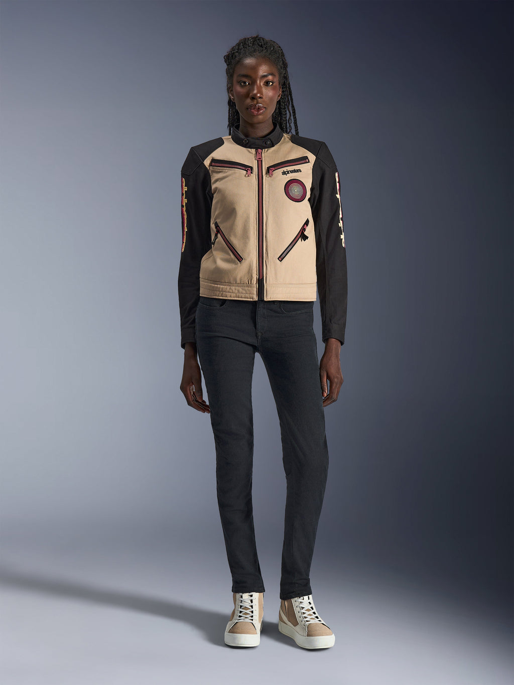 Chaqueta Alpinestars Stella Grace para mujer, chaqueta urbana para motocicleta, confección textil en color canela y negro, con cremalleras frontales y parche bordado Oscar en espiral, usada con pantalones negros y zapatillas de caña alta, equipamiento de conducción contemporáneo inspirado en la ropa de trabajo.