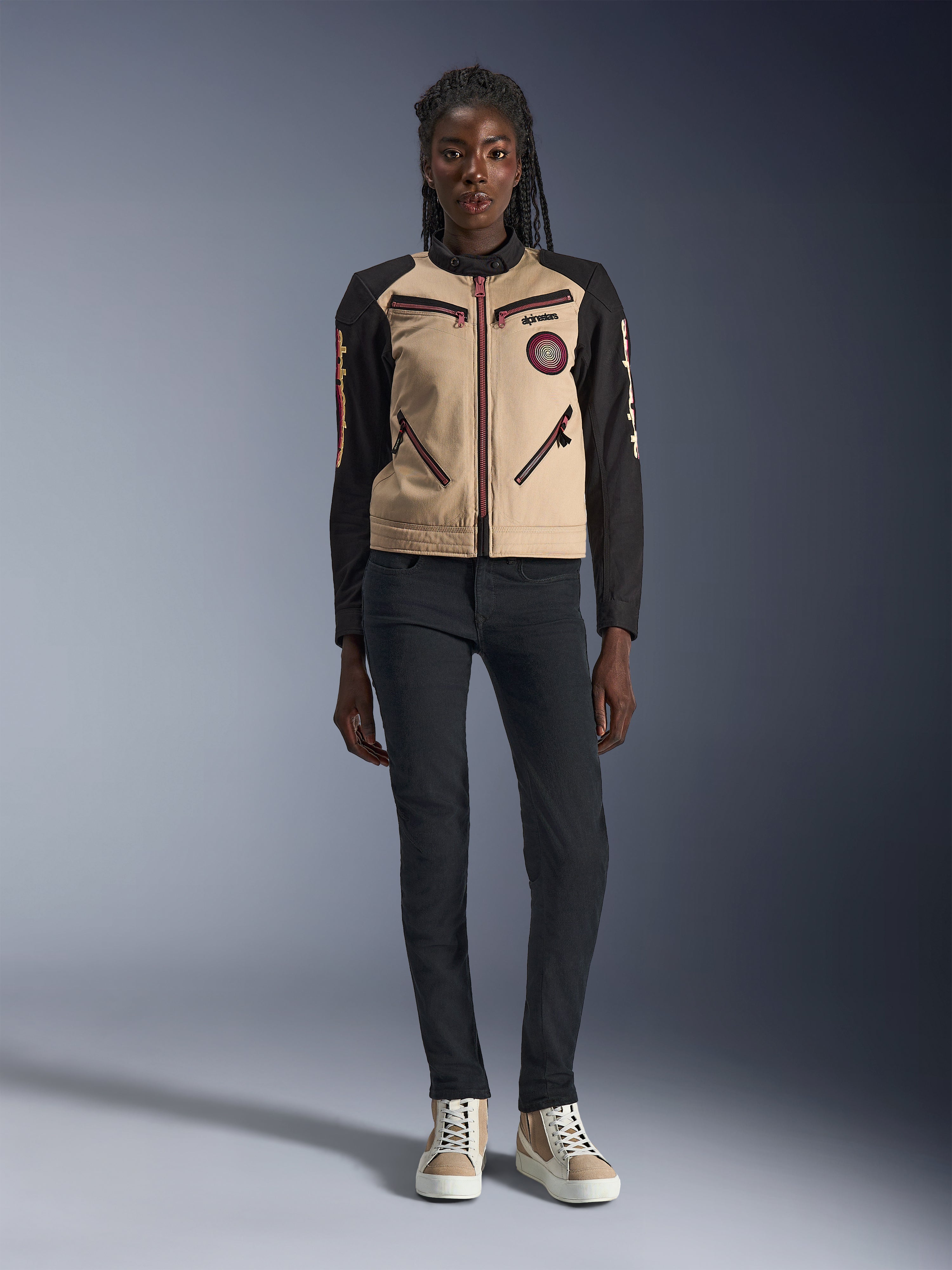 Chaqueta Alpinestars Stella Grace para mujer, chaqueta urbana para motocicleta, confección textil en color canela y negro, con cremalleras frontales y parche bordado Oscar en espiral, usada con pantalones negros y zapatillas de caña alta, equipamiento de conducción contemporáneo inspirado en la ropa de trabajo.