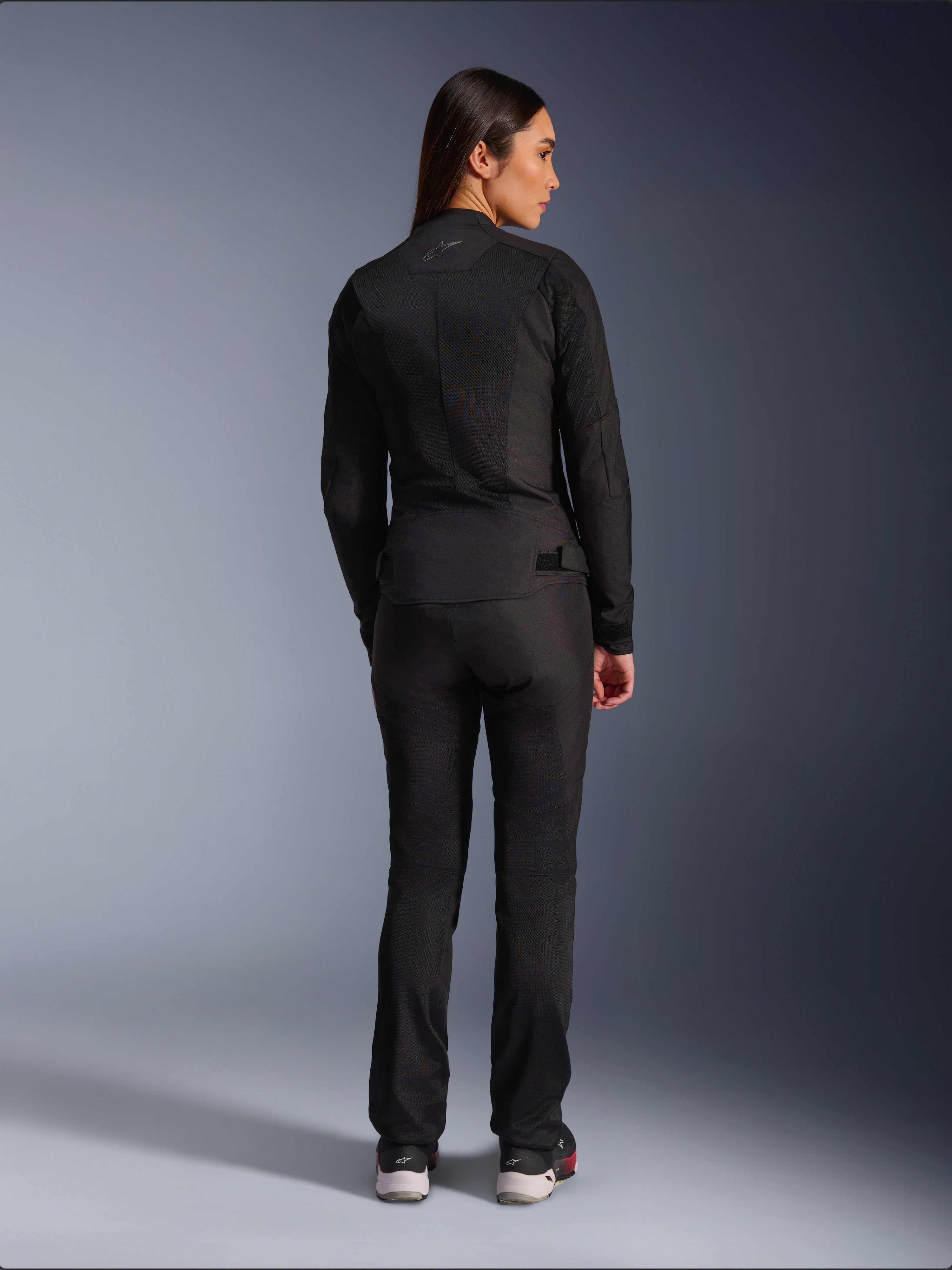 Chaqueta Alpinestars Stella C-1 Air, chaqueta de moto textil para mujer, negra, vista trasera mostrando la construcción de malla y un sutil logotipo en la parte superior de la espalda, combinada con pantalones de conducción negros a juego, estilo urbano contemporáneo