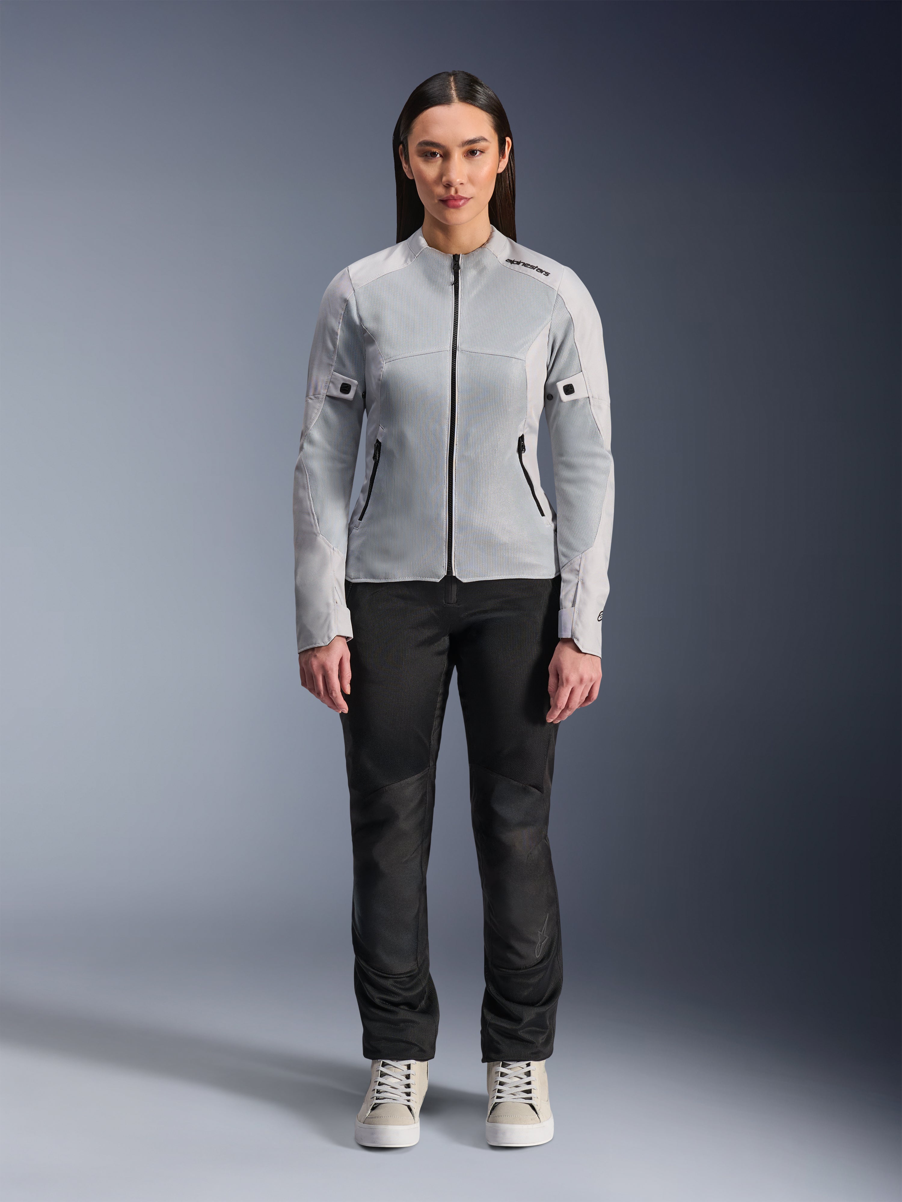 Mujer con chaqueta Alpinestars Stella C-1 Air en plateado, vista frontal, chaqueta de moto textil urbana con paneles de malla técnica, combinada con pantalones negros y zapatillas blancas, equipamiento de conducción contemporáneo
