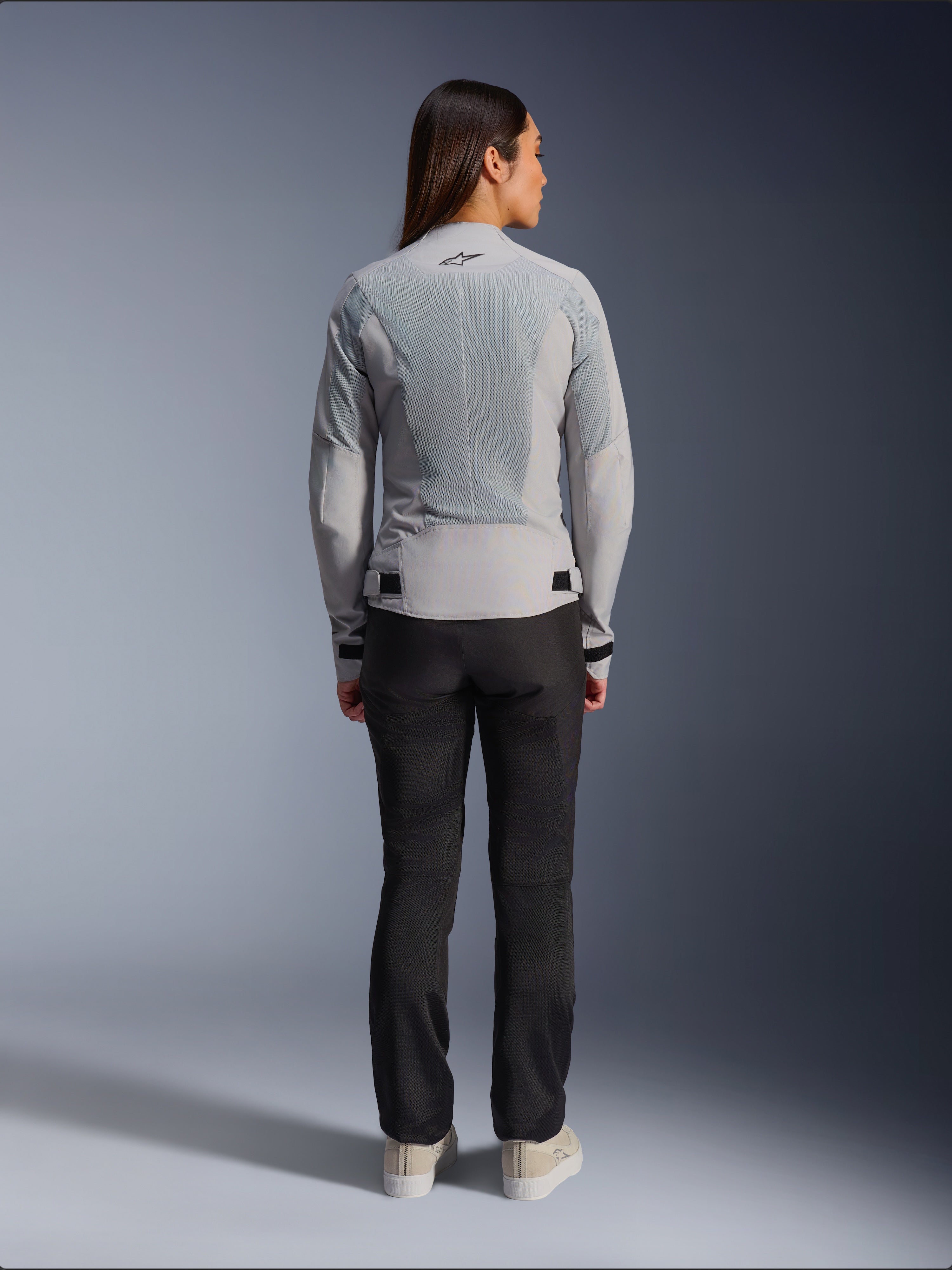 Mujer con la chaqueta Alpinestars Stella C-1 Air en color plateado, vista trasera, con grandes paneles de malla técnica para ventilación y un logotipo de estrella reflectante, combinada con pantalones de motociclista negros y zapatillas blancas, equipamiento de moto urbano contemporáneo