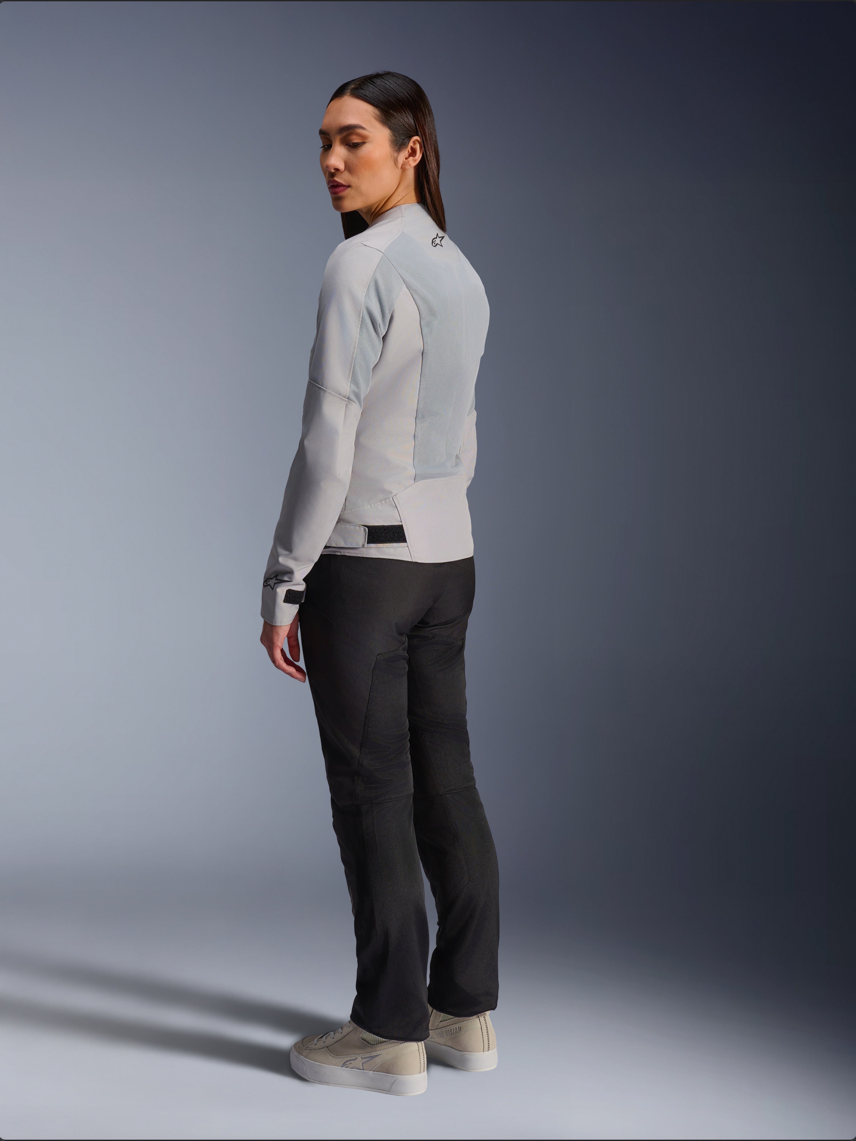Mujer con la chaqueta Alpinestars Stella C-1 Air en plateado, vista trasera y lateral que muestra los paneles de malla técnica y el logotipo reflectante en el hombro, combinada con pantalones de moto negros y zapatillas beige, estética de conducción urbana contemporánea