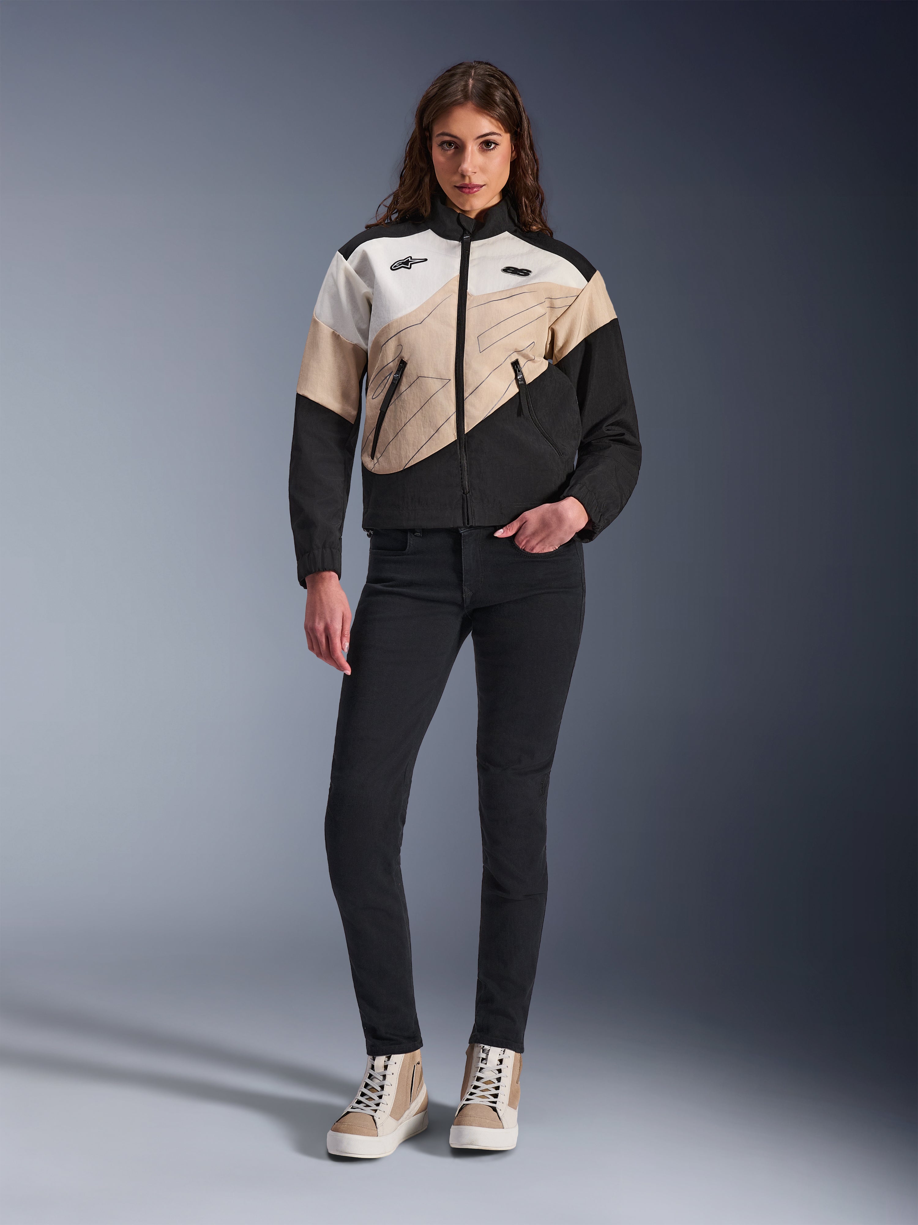 Chaqueta Alpinestars Stella Moshell para mujer, chaqueta urbana de motociclismo con un diseño asimétrico de bloques de color en negro, arena y blanco roto, combinada con pantalones negros de corte ajustado y zapatillas altas de color arena, estética streetwear moderna para motoristas.