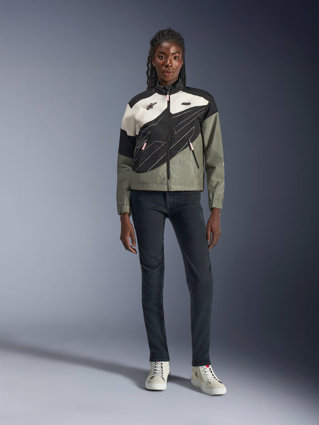 Chaqueta Alpinestars Stella Moshell para mujer, en color negro, gris y blanco roto con detalles en rosa, lucida por una modelo con vaqueros oscuros y zapatillas blancas, con diseño de cremallera asimétrica y gráficos geométricos llamativos, equipamiento urbano contemporáneo