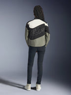 Vista trasera de una modelo con la chaqueta Alpinestars Stella Moshell en negro, gris y blanco roto, con un diseño asimétrico y un gran logotipo perfilado en la espalda, combinada con vaqueros oscuros ajustados y zapatillas blancas, ropa de moto para mujer de estilo urbano.