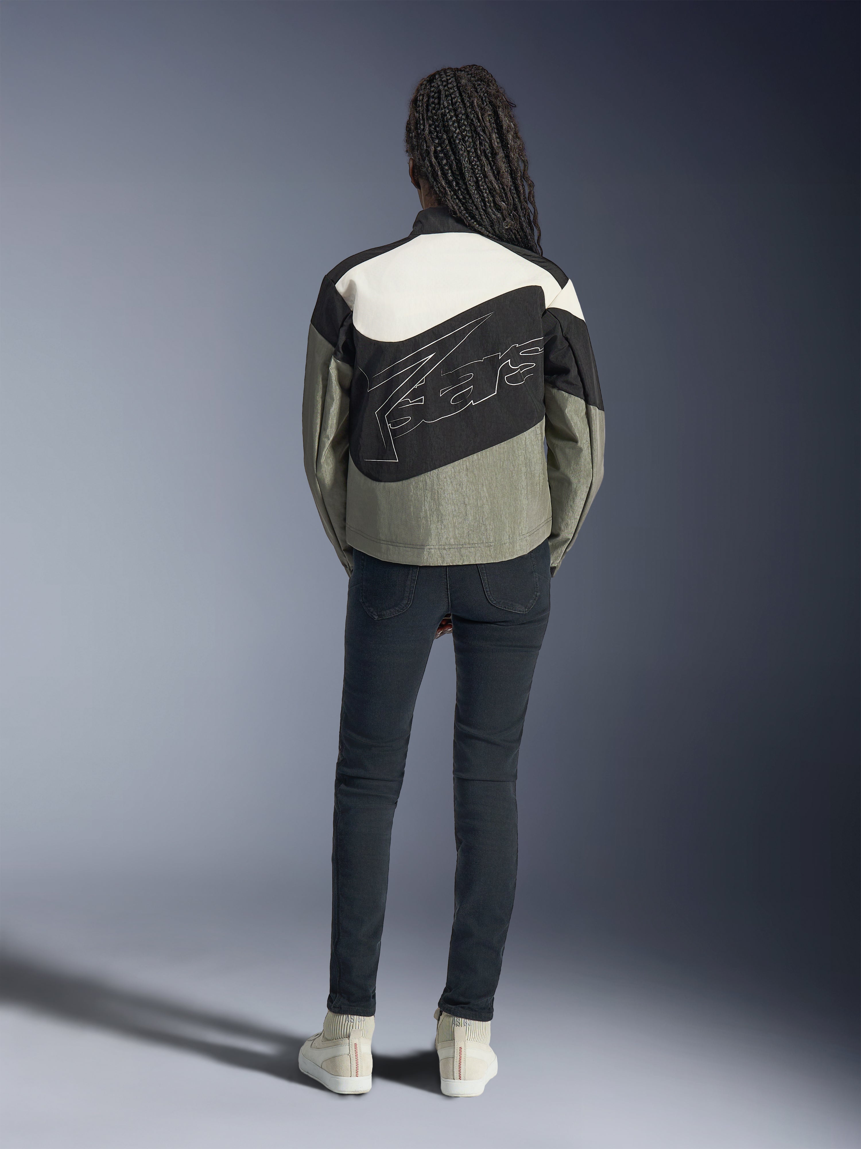 Vista trasera de una modelo con la chaqueta Alpinestars Stella Moshell en negro, gris y blanco roto, con un diseño asimétrico y un gran logotipo perfilado en la espalda, combinada con vaqueros oscuros ajustados y zapatillas blancas, ropa de moto para mujer de estilo urbano.