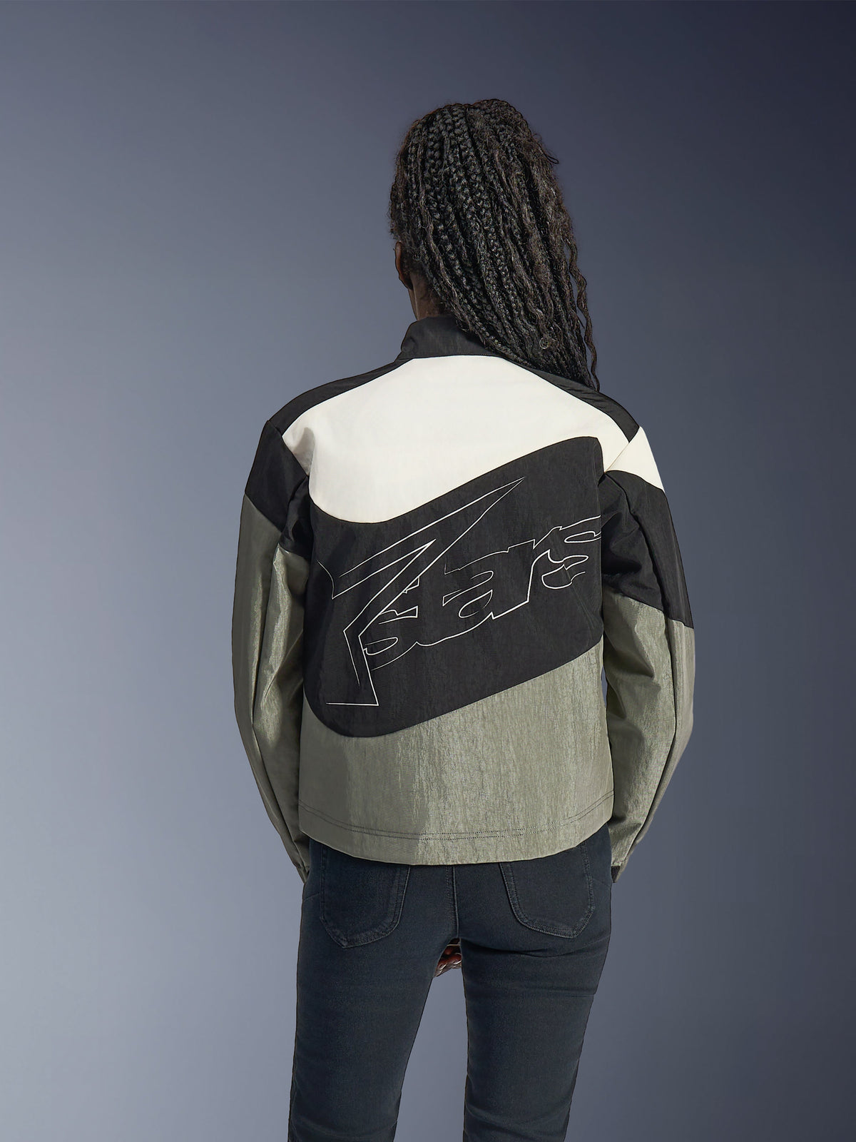 Mujer de espaldas con la chaqueta Alpinestars Stella Moshell, chaqueta de moto urbana en negro, gris y blanco roto, con un gran logotipo blanco perfilado en la espalda, combinada con pantalones negros, equipamiento de conducción contemporáneo inspirado en el estilo streetwear.