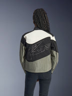 Mujer de espaldas con la chaqueta Alpinestars Stella Moshell, chaqueta de moto urbana en negro, gris y blanco roto, con un gran logotipo blanco perfilado en la espalda, combinada con pantalones negros, equipamiento de conducción contemporáneo inspirado en el estilo streetwear.