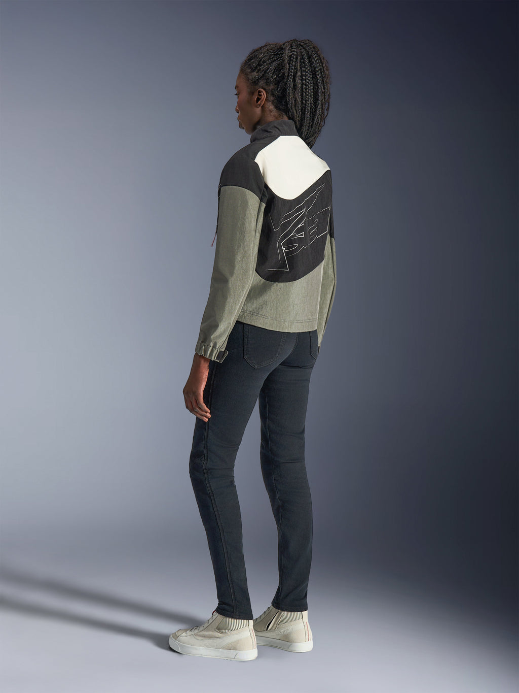 Mujer con la chaqueta Alpinestars Stella Moshell, chaqueta urbana de moto, en colores negro, gris y blanco roto, vista trasera con diseño de paneles asimétricos y un gráfico grande en la espalda, combinada con pantalones negros ajustados y zapatillas blanco roto, equipamiento contemporáneo de moto urbana