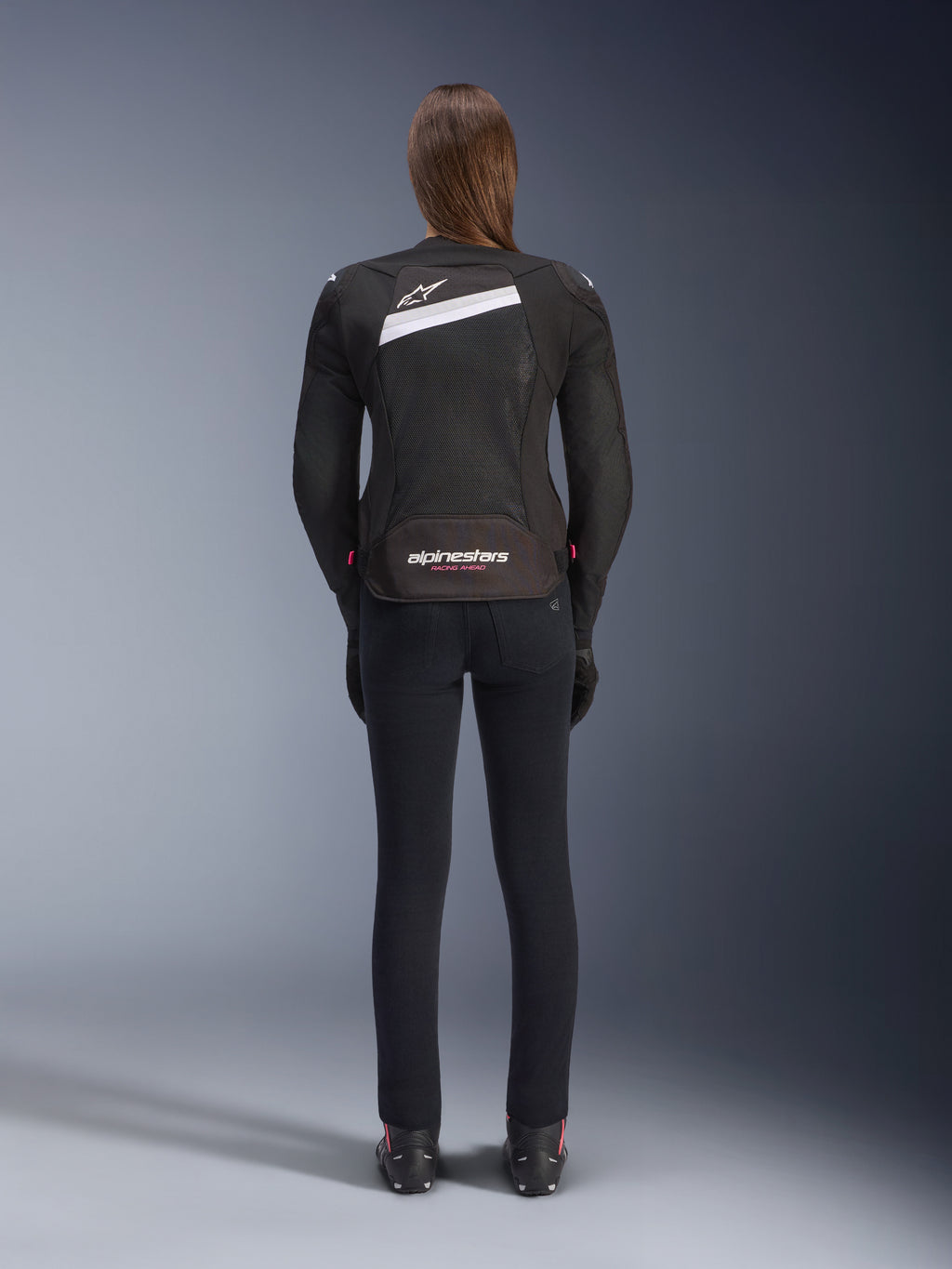 Chaqueta Alpinestars Stella T-GP Plus R V4 Airflow, negra y blanca, vista trasera que muestra paneles de ventilación de malla extensiva y la marca Alpinestars, usada por una motociclista con pantalones de moto negros de ajuste ceñido y botas, equipamiento textil deportivo para mujer