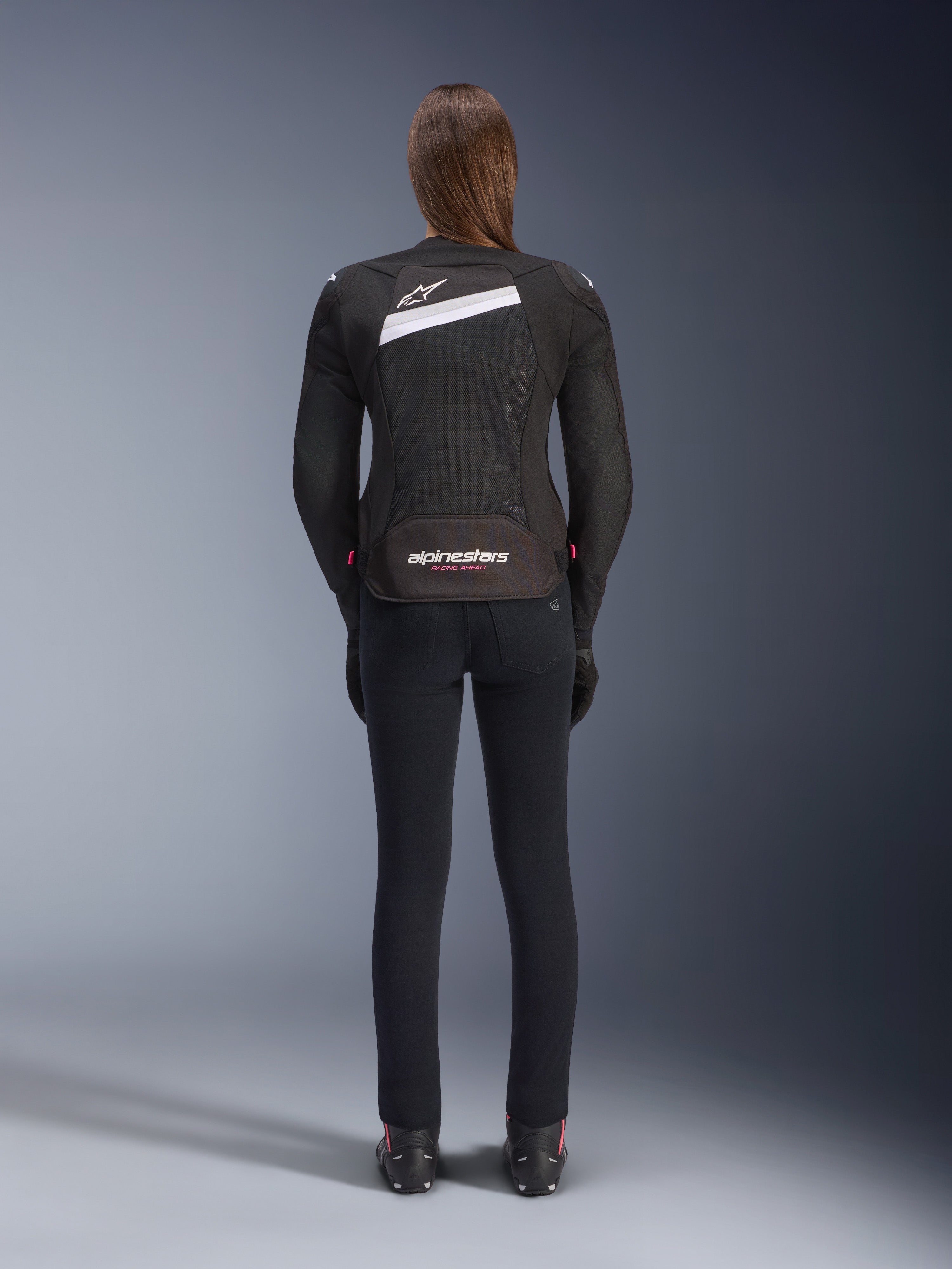Chaqueta Alpinestars Stella T-GP Plus R V4 Airflow, negra y blanca, vista trasera que muestra paneles de ventilación de malla extensiva y la marca Alpinestars, usada por una motociclista con pantalones de moto negros de ajuste ceñido y botas, equipamiento textil deportivo para mujer