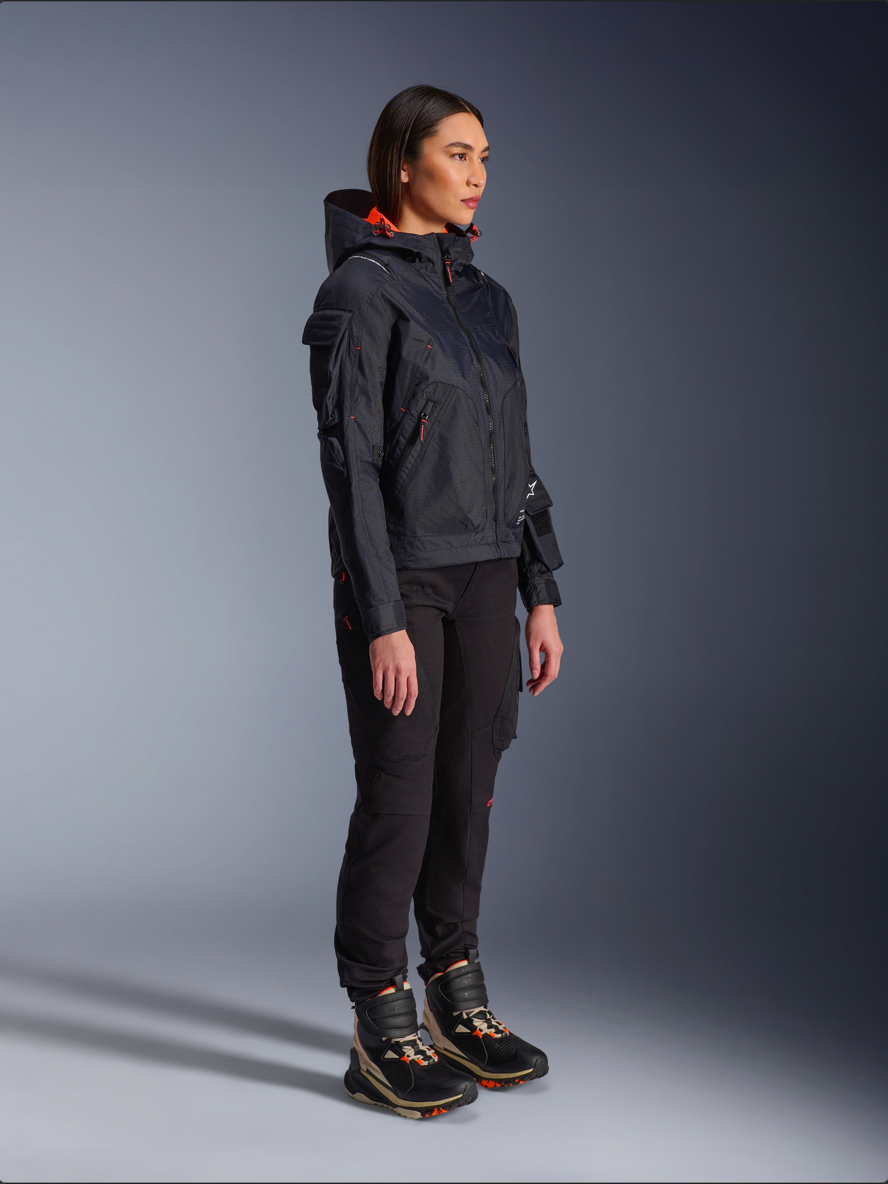Pantalones Alpinestars Actea para mujer en negro, corte regular con bolsillos cargo, combinados con una chaqueta Molly Air negra con detalles naranjas y calzado de conducción de caña alta, ropa de moto urbana contemporánea para mujer