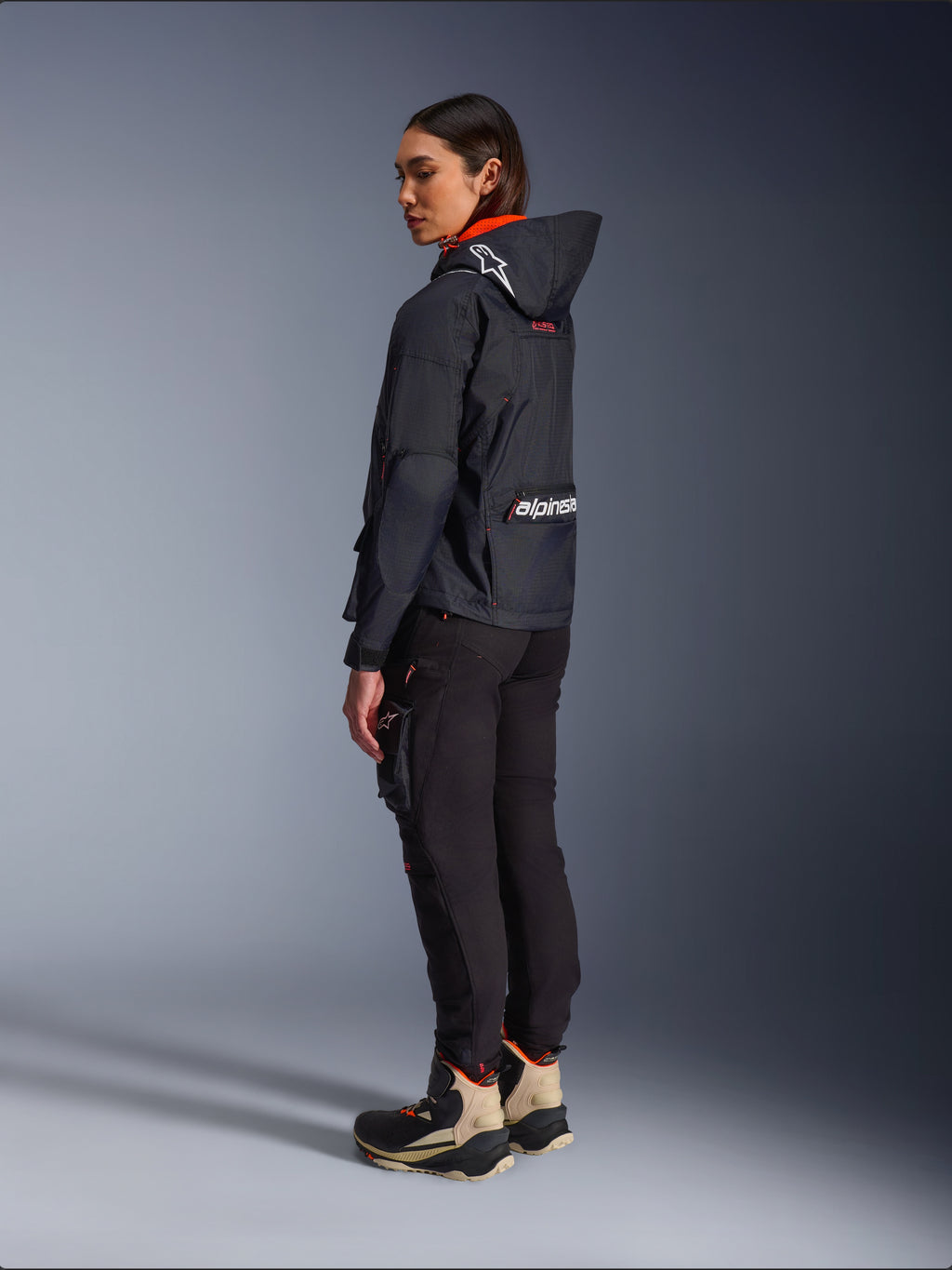 Pantalones Alpinestars Actea para mujer, negros, modernos pantalones de moto con corte ajustado y bolsillo cargo en el muslo, mostrados desde una perspectiva lateral trasera en una modelo que viste una chaqueta oscura con capucha y botas de conducción, estilo de conducción urbana.