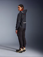 Pantalones Alpinestars Actea para mujer, negros, modernos pantalones de moto con corte ajustado y bolsillo cargo en el muslo, mostrados desde una perspectiva lateral trasera en una modelo que viste una chaqueta oscura con capucha y botas de conducción, estilo de conducción urbana.