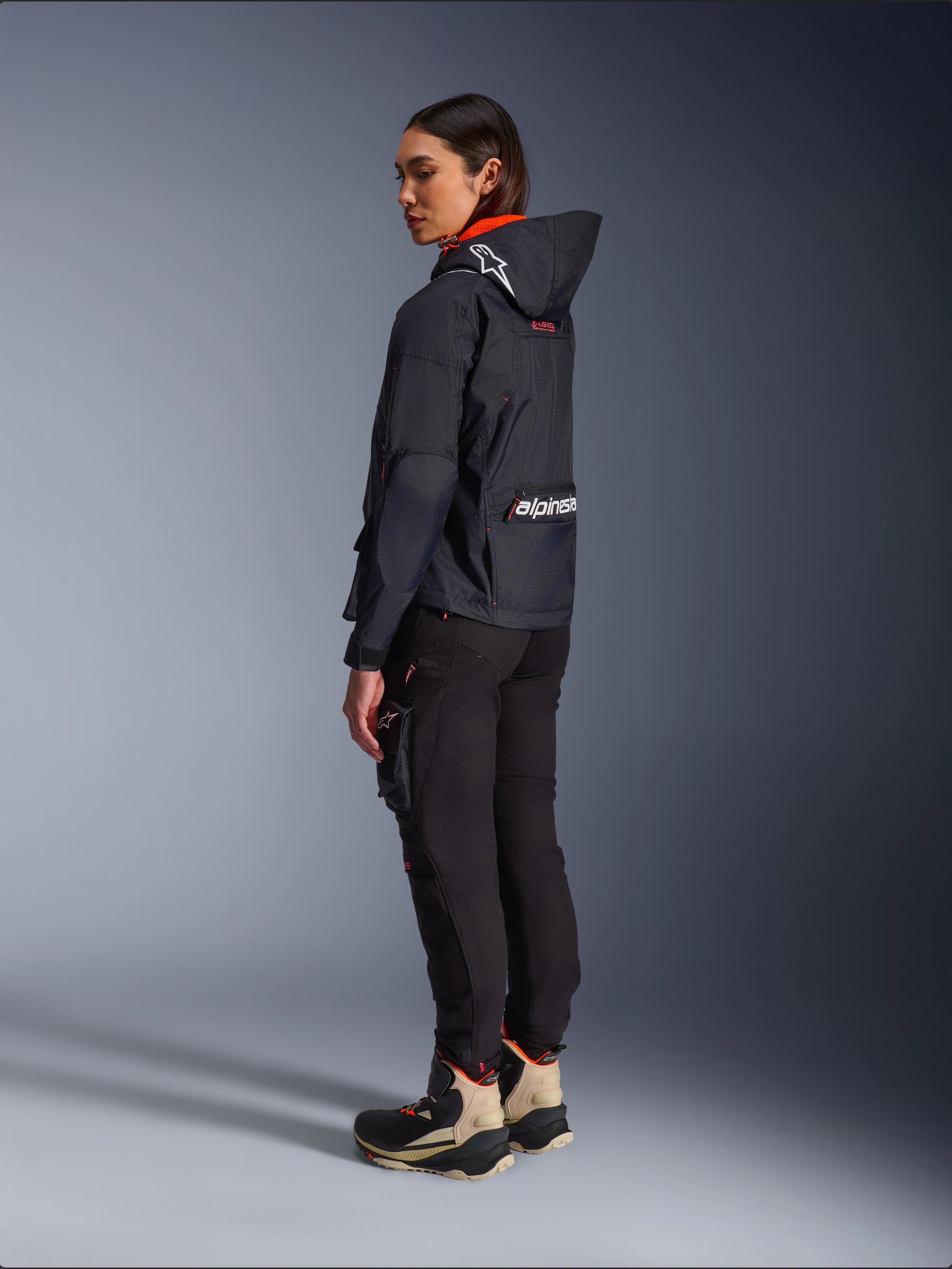 Pantalones Alpinestars Actea para mujer, negros, modernos pantalones de moto con corte ajustado y bolsillo cargo en el muslo, mostrados desde una perspectiva lateral trasera en una modelo que viste una chaqueta oscura con capucha y botas de conducción, estilo de conducción urbana.