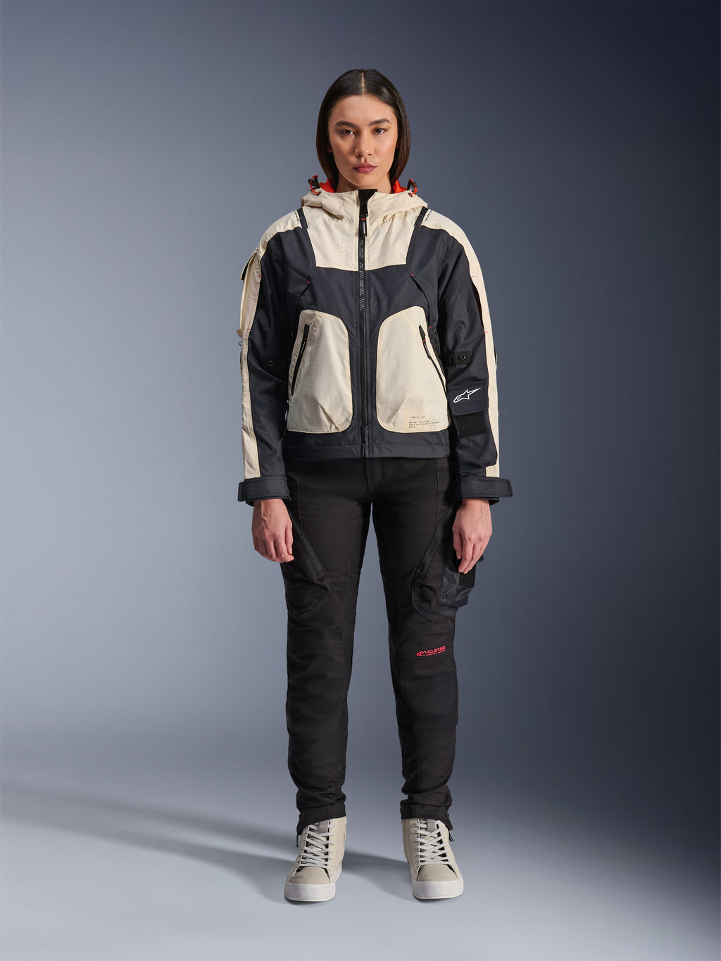 Female model wearing the Alpinestars Molly Air Jacket in Black Almond, featuring a color-blocked design with cream and black panels and a red-lined hood, paired with black motorcycle pants and cream sneakers, studio shot of urban contemporary riding gear.
