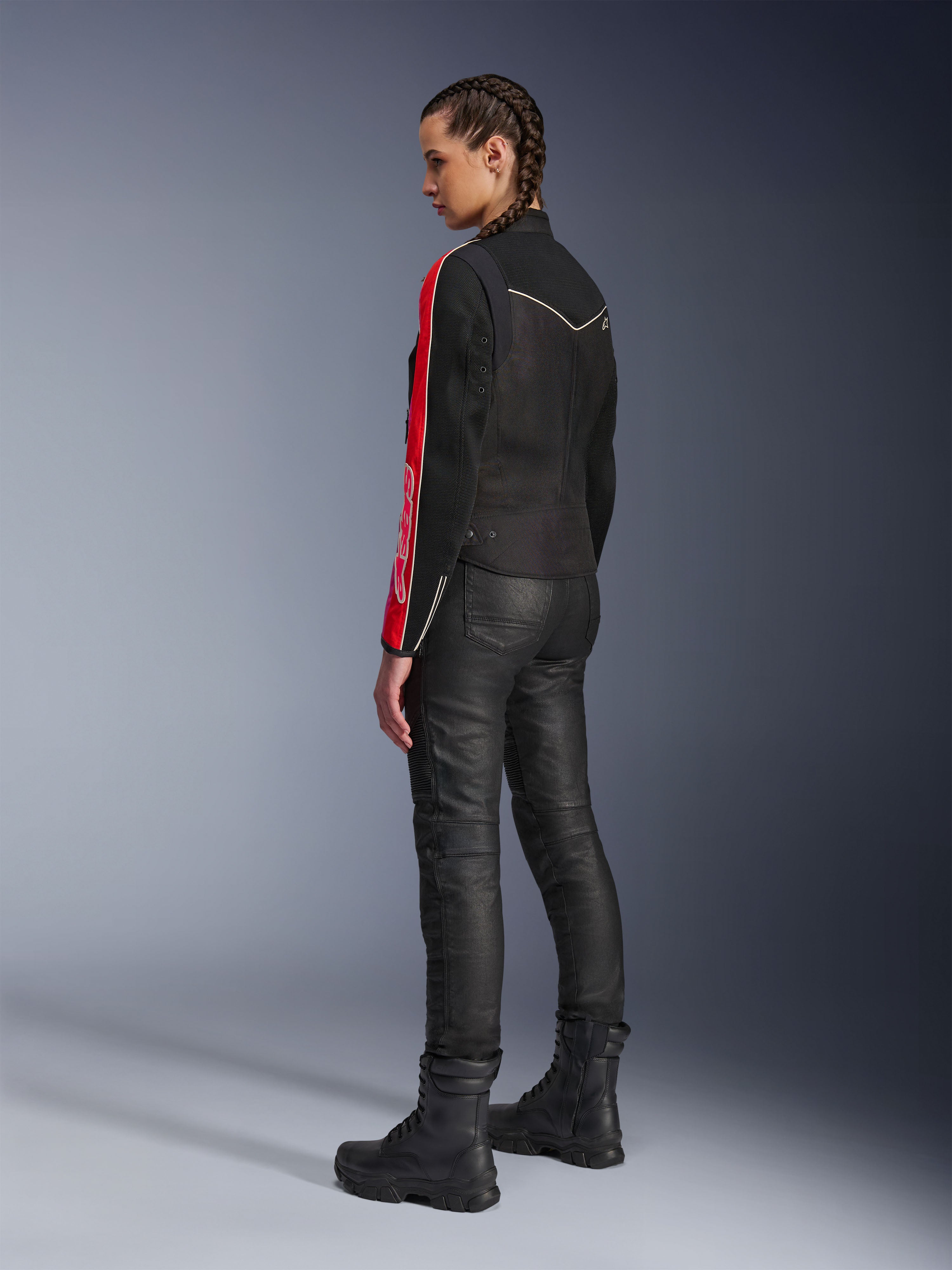 Mujer con la chaqueta Alpinestars Stella T-Dyno Air en color Negro Rojo Brillante Fucsia, combinada con pantalones de motociclista negros y botas negras, vista de perfil que resalta los gráficos rojos y fucsia de las mangas y el ajuste de motociclista entallado.
