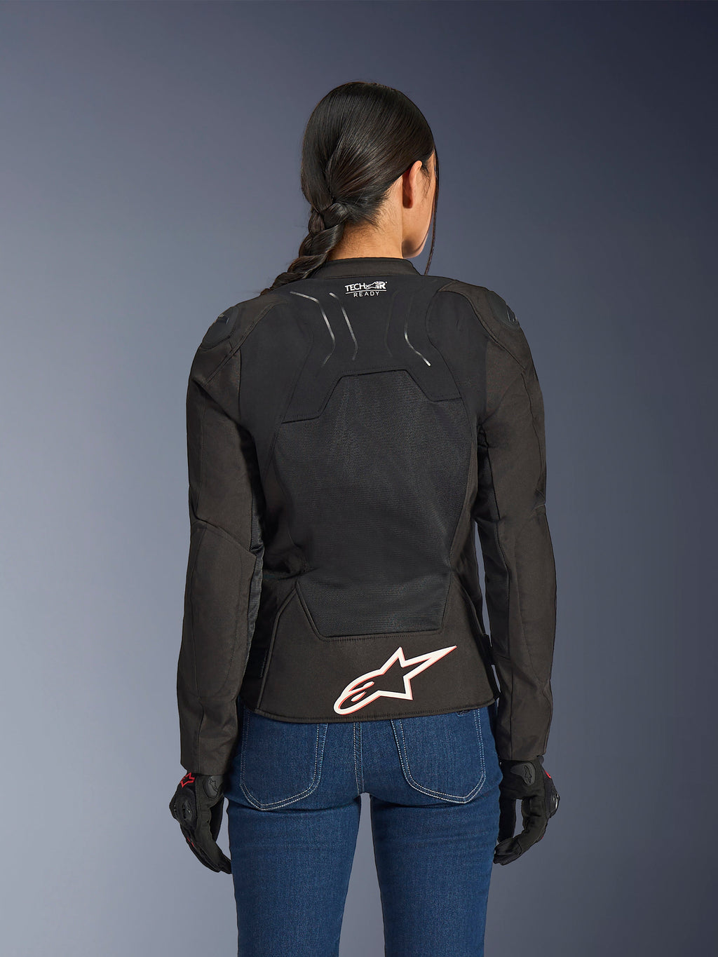 Chaqueta Alpinestars Stella SMX Air, vista trasera en modelo femenina, textil negro con paneles de malla de alto flujo, marca 'TECH-AIR READY' en la parte superior de la espalda, logotipo grande de Alpinestars en blanco con contorno rojo en la parte inferior de la espalda, combinada con vaqueros azules y guantes de conducción negros