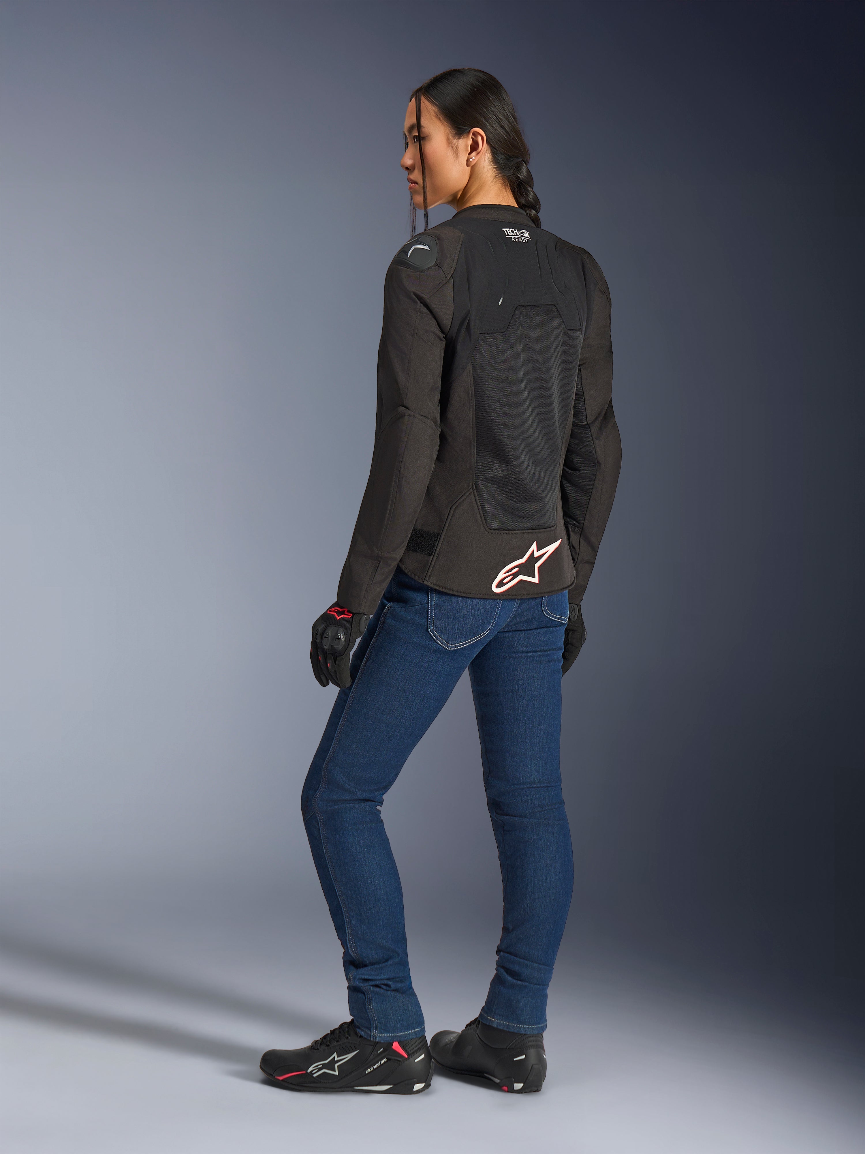 Chaqueta Alpinestars Stella SMX Air, negra con logotipo blanco y rojo, lucida por una motociclista en una vista trasera de tres cuartos, combinada con vaqueros de moto azules y calzado de conducción negro, con construcción de malla técnica para conducción deportiva en climas cálidos.